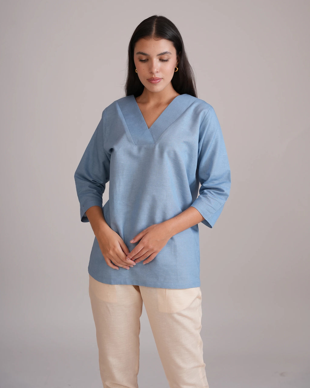 A woman wearing a light blue V-neck scrub top with three-quarter sleeves and beige trousers, standing against a plain light gray studio background.