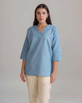 A woman modeling a light blue V-neck tunic top with three-quarter sleeves, paired with beige trousers, against a plain light gray studio background.