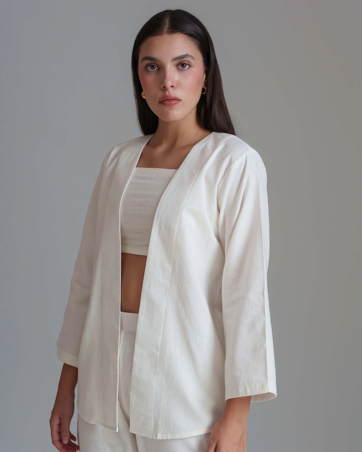 A woman modeling a cream-colored three-quarter sleeve open-front jacket over a matching cream crop top and trousers, set against a solid gray studio background.