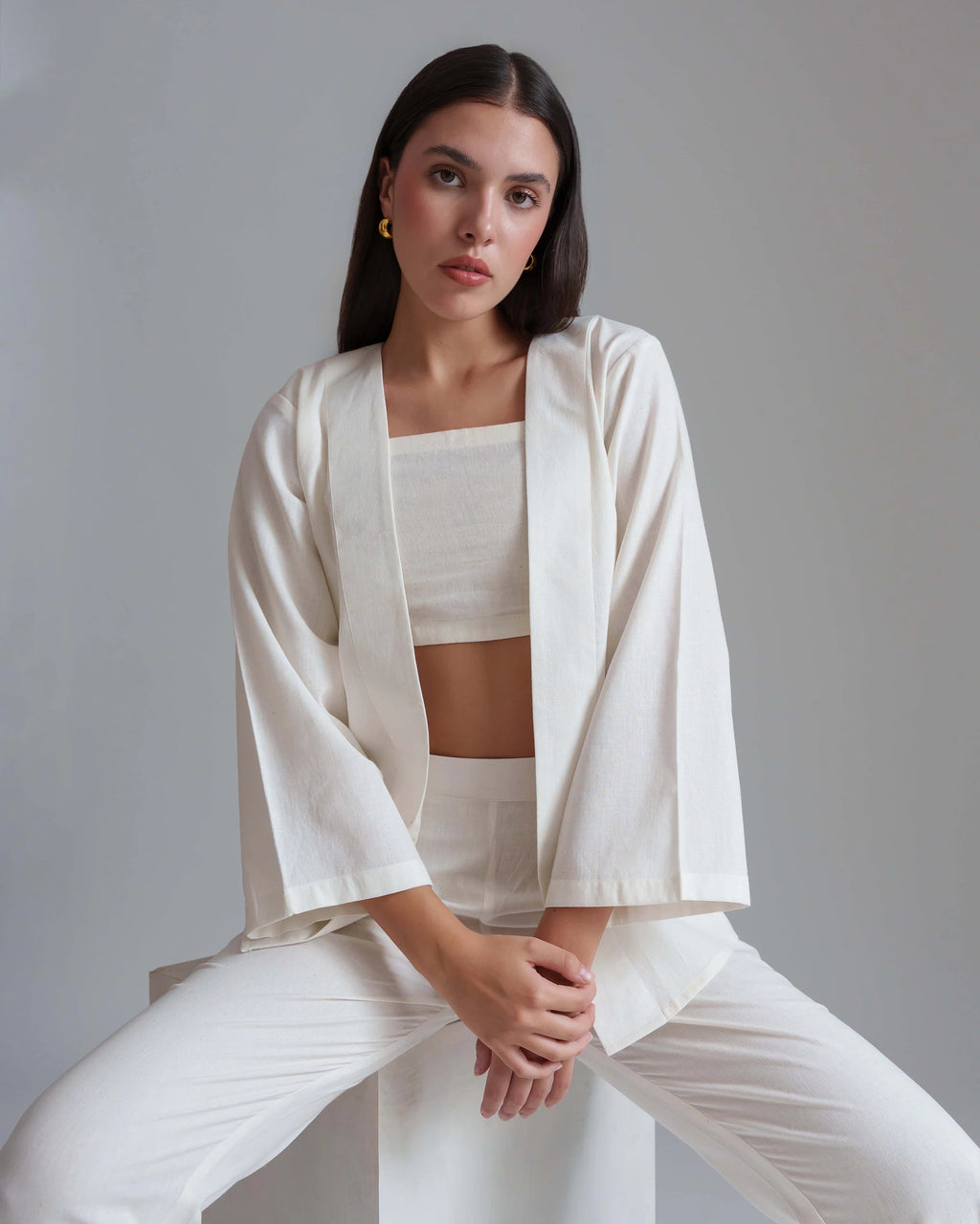 A woman seated on a white block wearing a matching white lounge set consisting of a cropped tank top, open-front long-sleeve jacket, and wide-leg pants against a plain gray studio background.