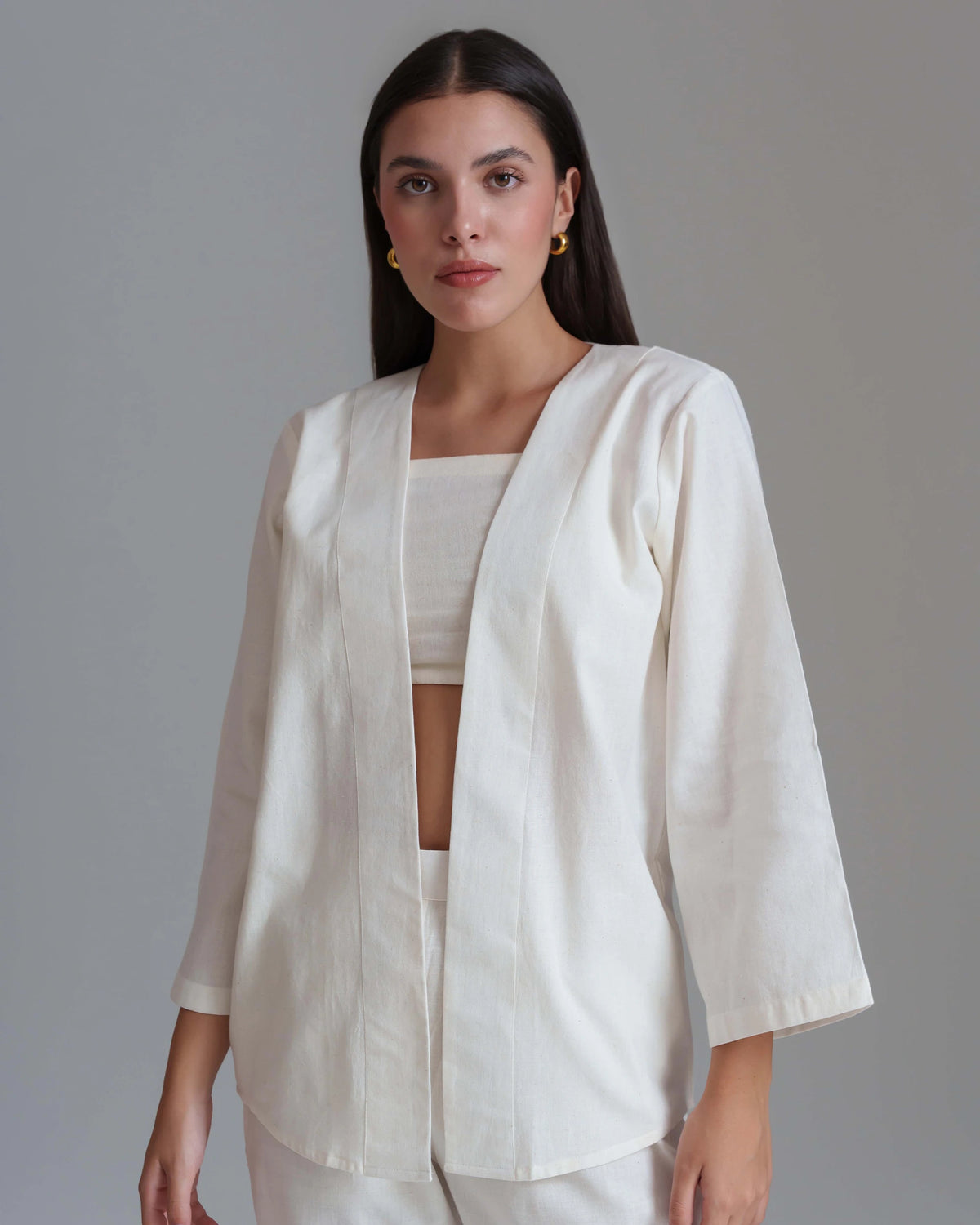 A woman modeling a white three-quarter sleeve open-front jacket over a matching white crop top and white pants, photographed against a plain gray studio background.