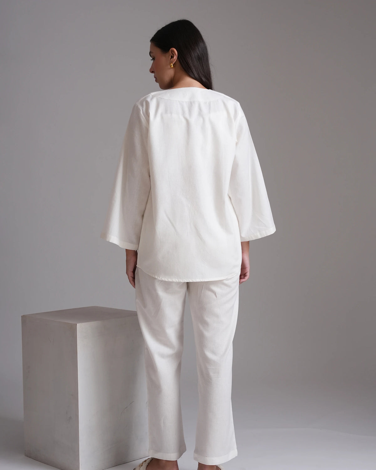 A woman stands with her back to the camera wearing a loose white two-piece outfit consisting of a short-sleeved top and matching wide-leg pants, posed next to a light gray concrete block against a neutral gray background.