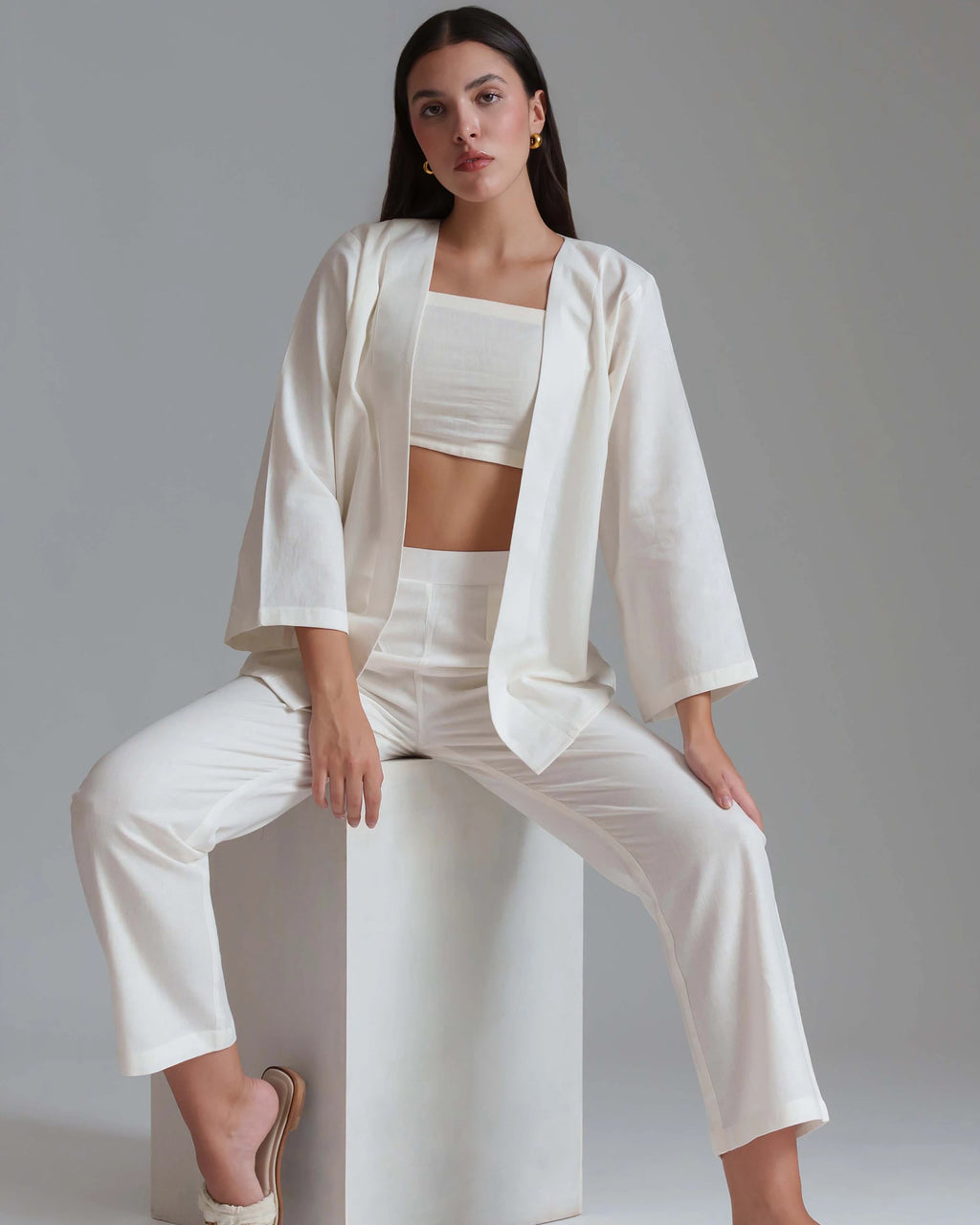 A woman models a matching white three-piece clothing set consisting of a cropped tank top, wide-sleeve blazer, and high-waisted cropped trousers, seated on a white block against a neutral gray background.