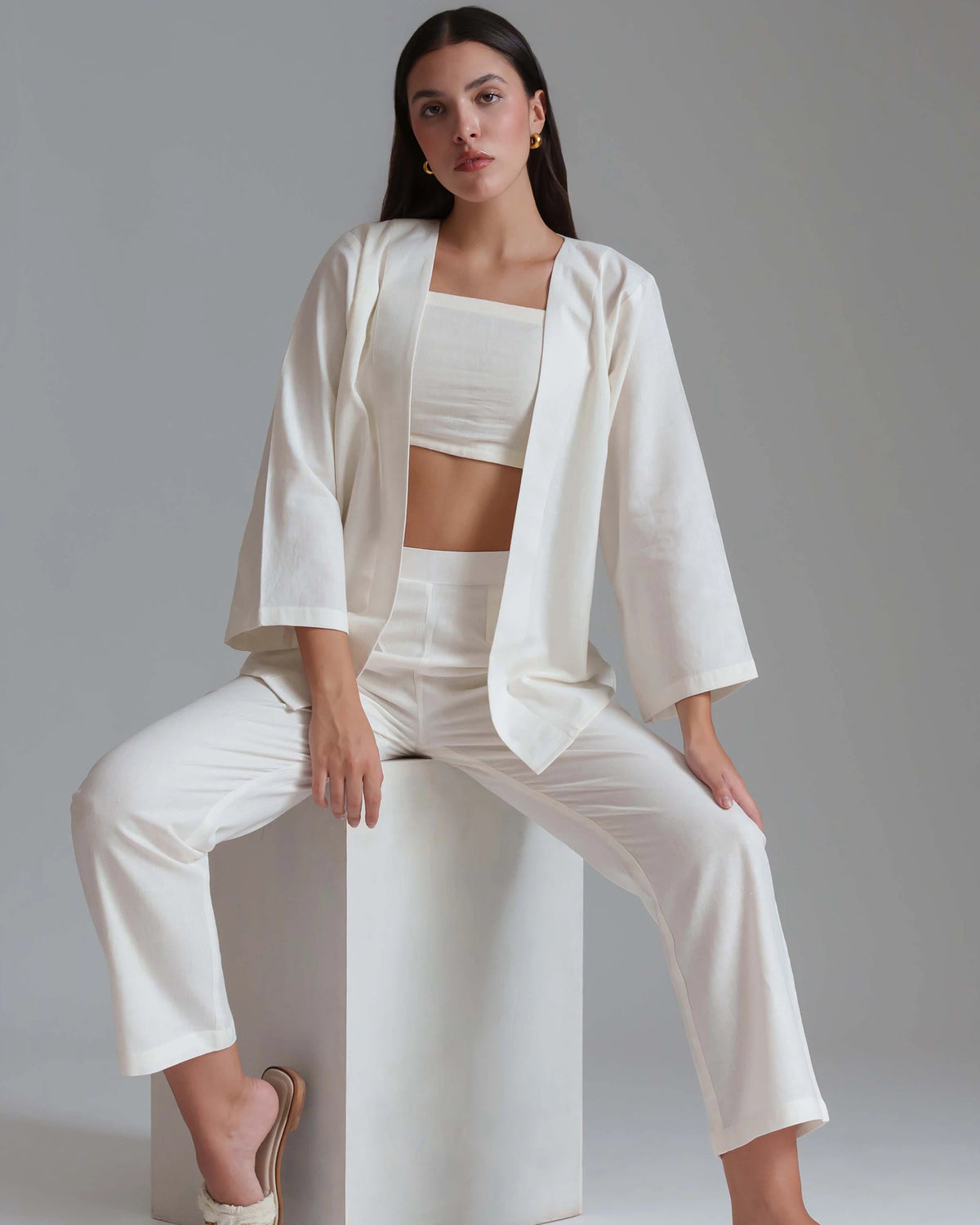 A woman models a matching white three-piece clothing set consisting of a cropped tank top, wide-sleeve blazer, and high-waisted cropped trousers, seated on a white block against a neutral gray background.
