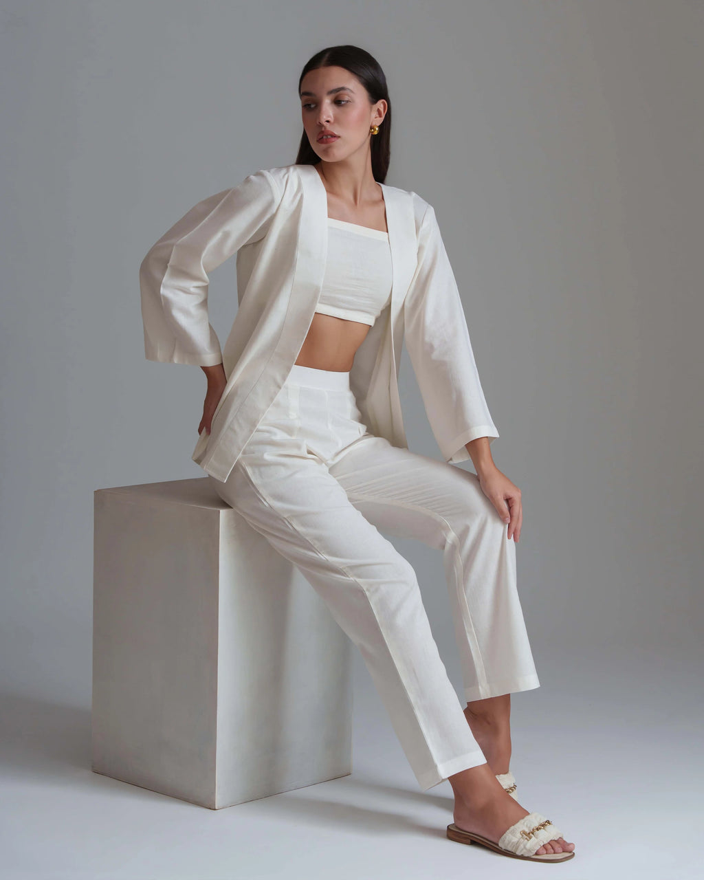 A model wearing a white three-piece clothing set consisting of a cropped tank top, matching trousers, and a draped open jacket, sitting on a white cube pedestal against a neutral gray background.