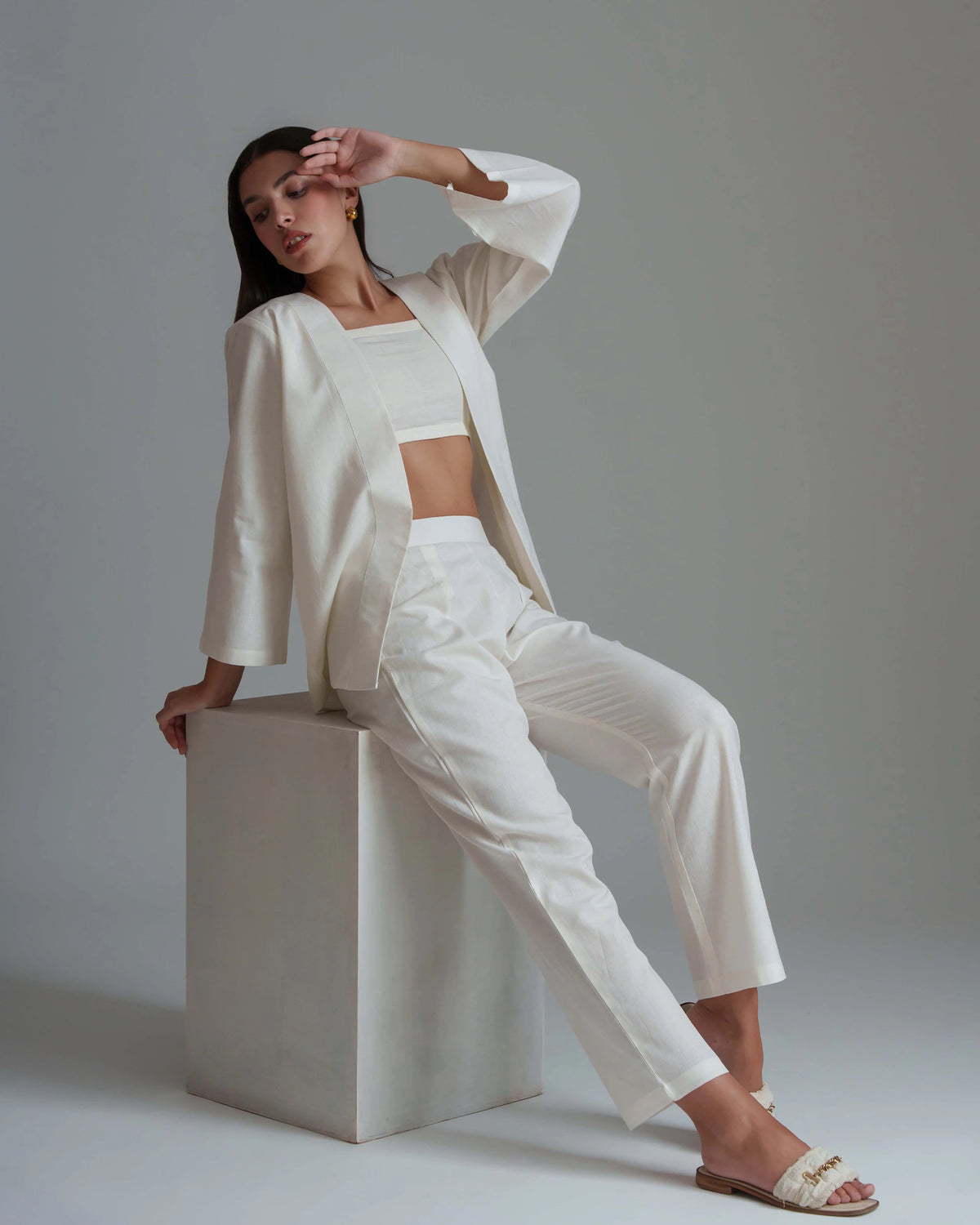 A model wearing a white three-piece suit consisting of a cropped blazer, a matching crop top, and high-waisted cropped trousers, sitting on a white cube pedestal against a neutral gray background.