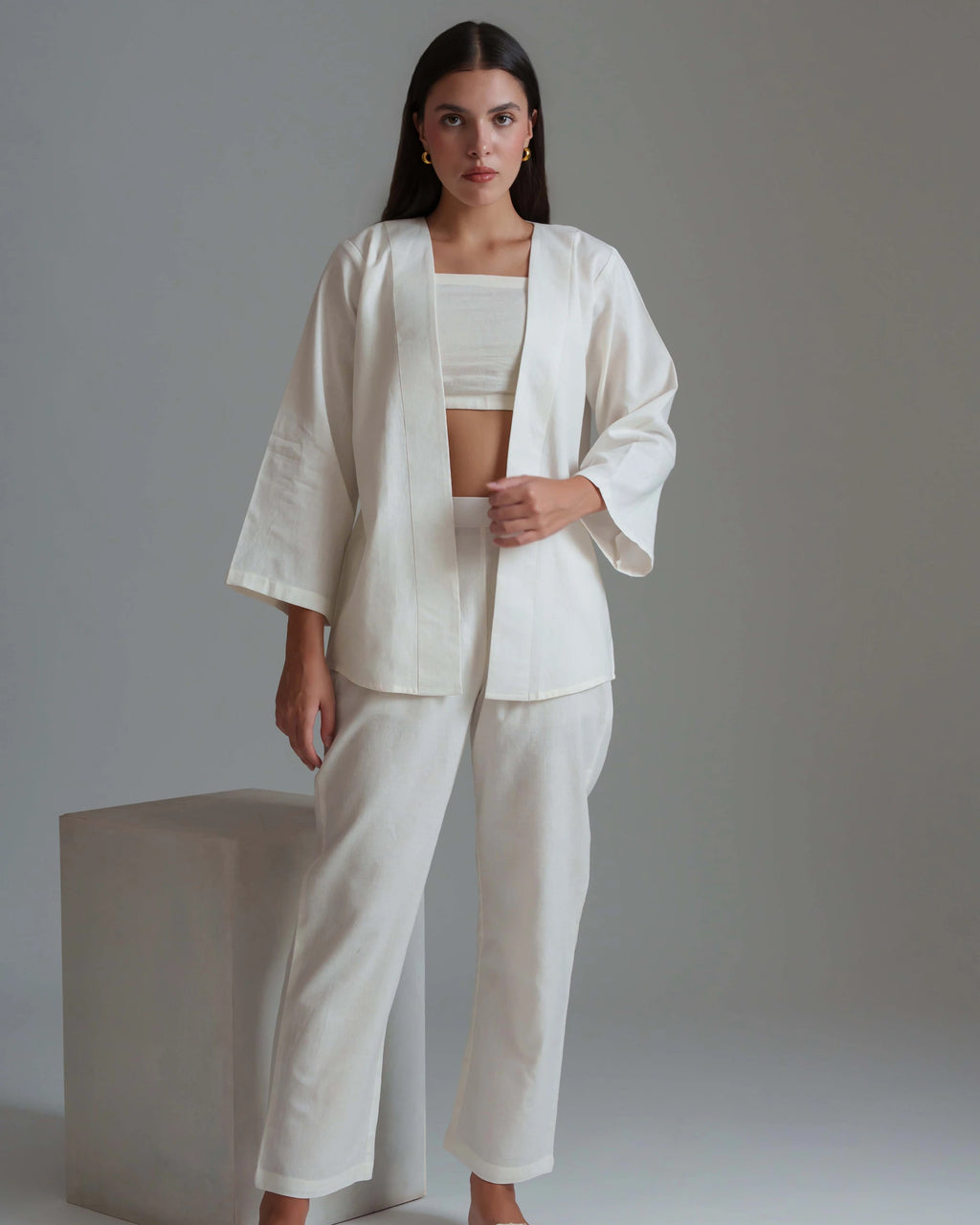A woman models a matching white three-piece linen suit consisting of a cropped tank top, a lightweight open-front jacket with wide sleeves, and wide-leg trousers, set against a neutral gray studio background.