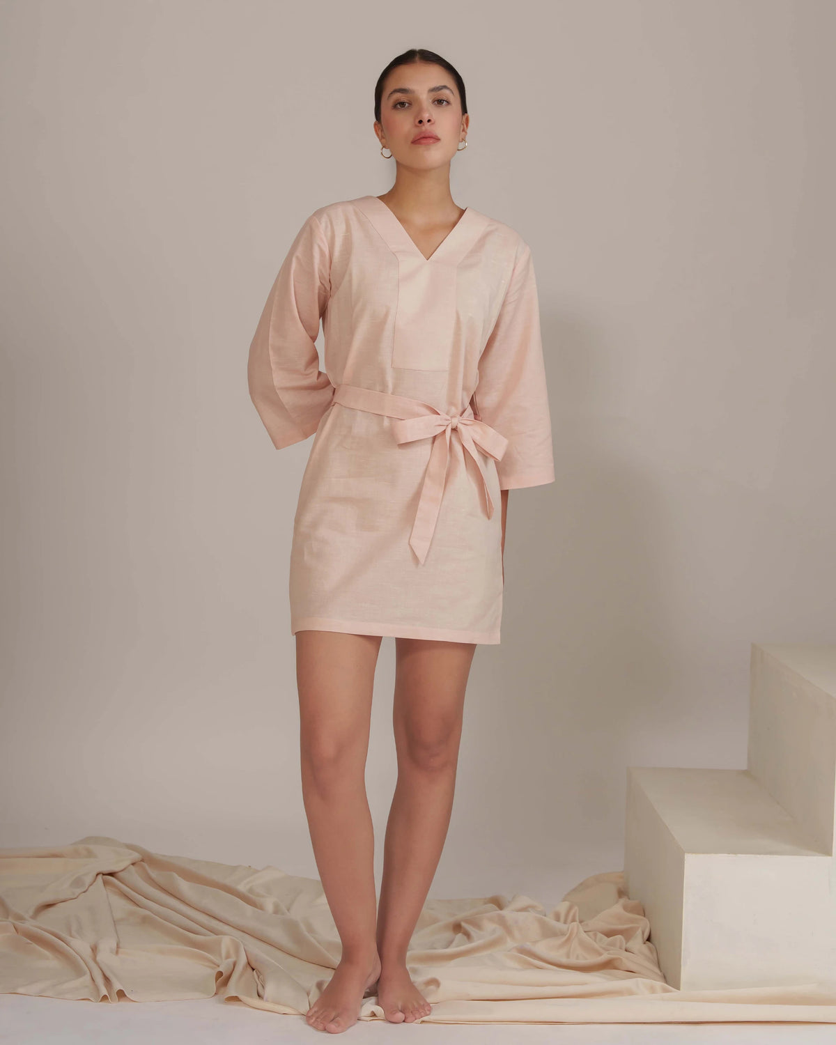 A woman stands barefoot in a studio wearing a light blush pink wrap robe dress with three-quarter sleeves and a tied waist. The background is a neutral warm beige with draped fabric on the floor and a white geometric block on the right.