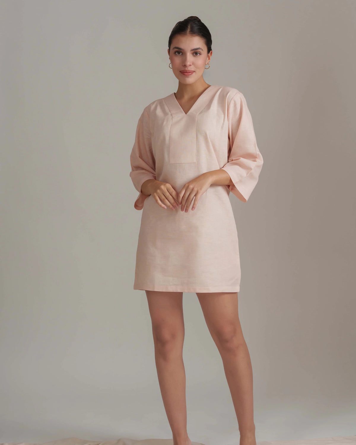A woman modeling a light blush pink linen tunic dress with three-quarter sleeves and a V-neckline, standing against a neutral beige studio backdrop.