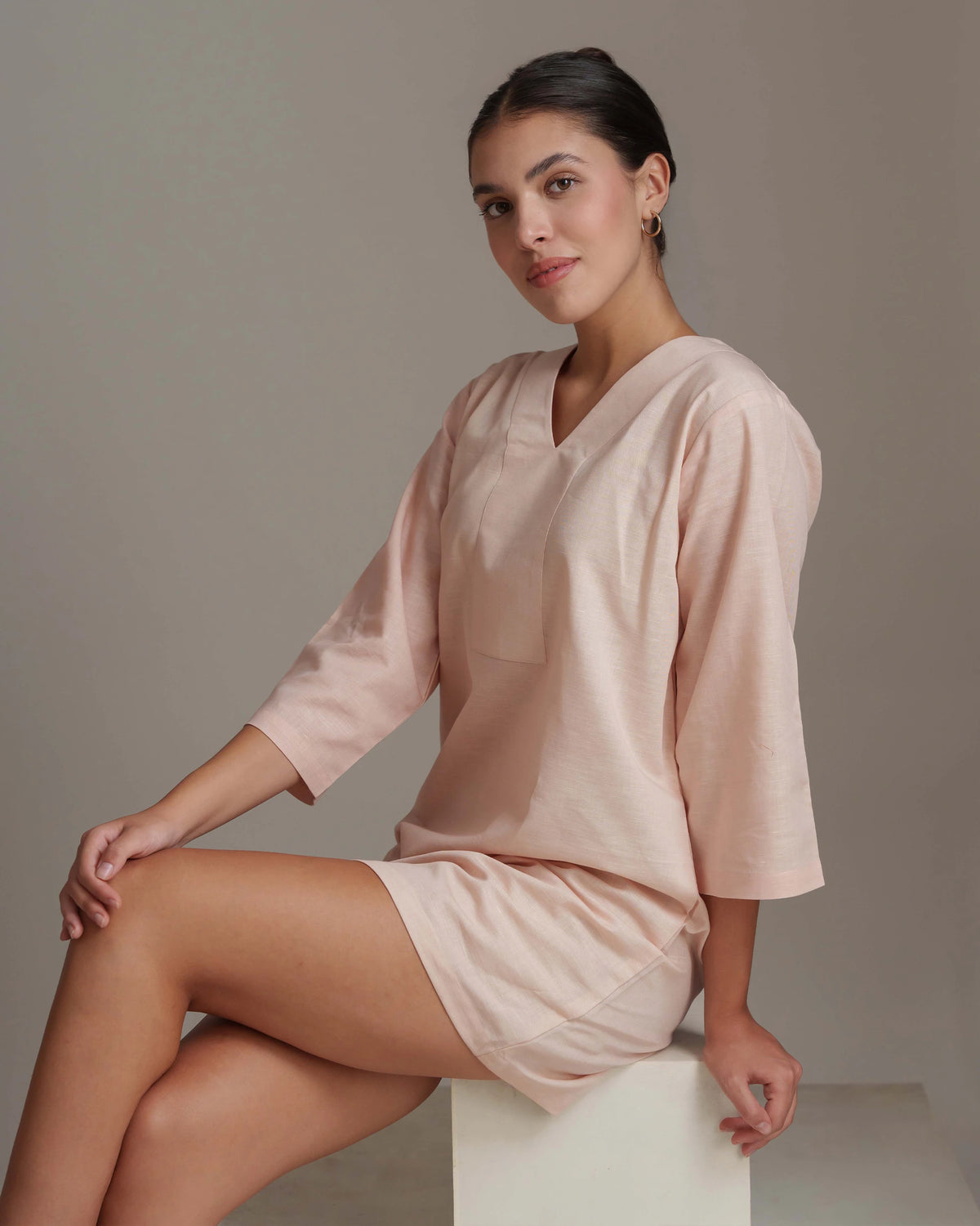 A woman wearing a light blush pink lounge set consisting of a short-sleeve V-neck top and matching shorts, sitting on a white block against a neutral gray studio background.