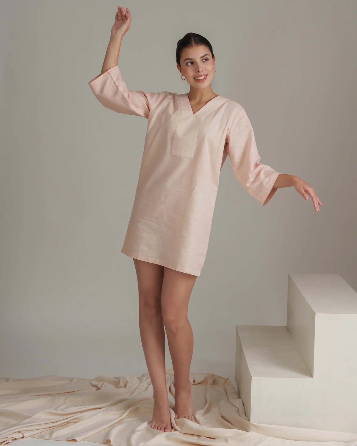 A woman modeling a light blush pink linen tunic dress with three-quarter sleeves and a V-neckline, standing in a studio setting with draped fabric and white geometric steps.