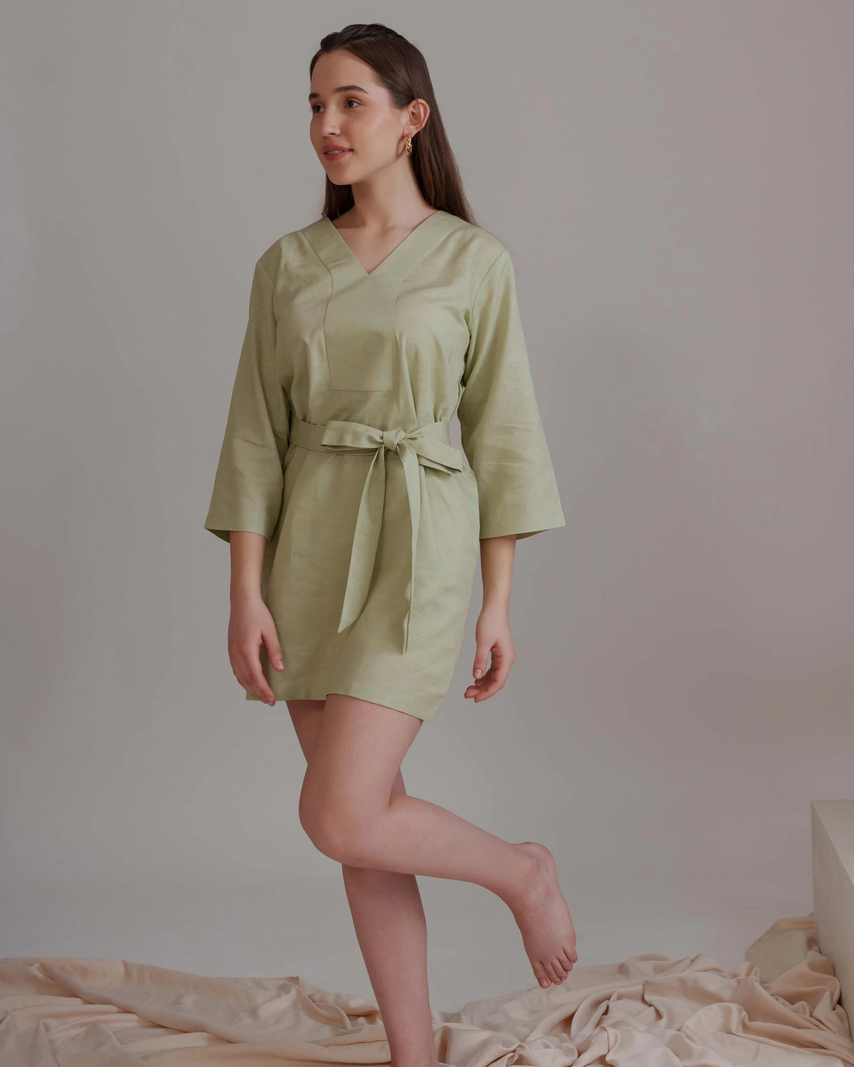 A woman modeling a light sage green kimono-style mini dress with three-quarter sleeves and a matching waist tie, standing against a neutral studio backdrop.