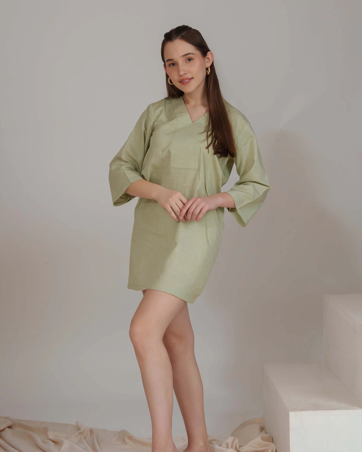 A woman modeling a light sage green short kimono-style dress with three-quarter sleeves and a V-neckline, standing against a neutral studio backdrop.
