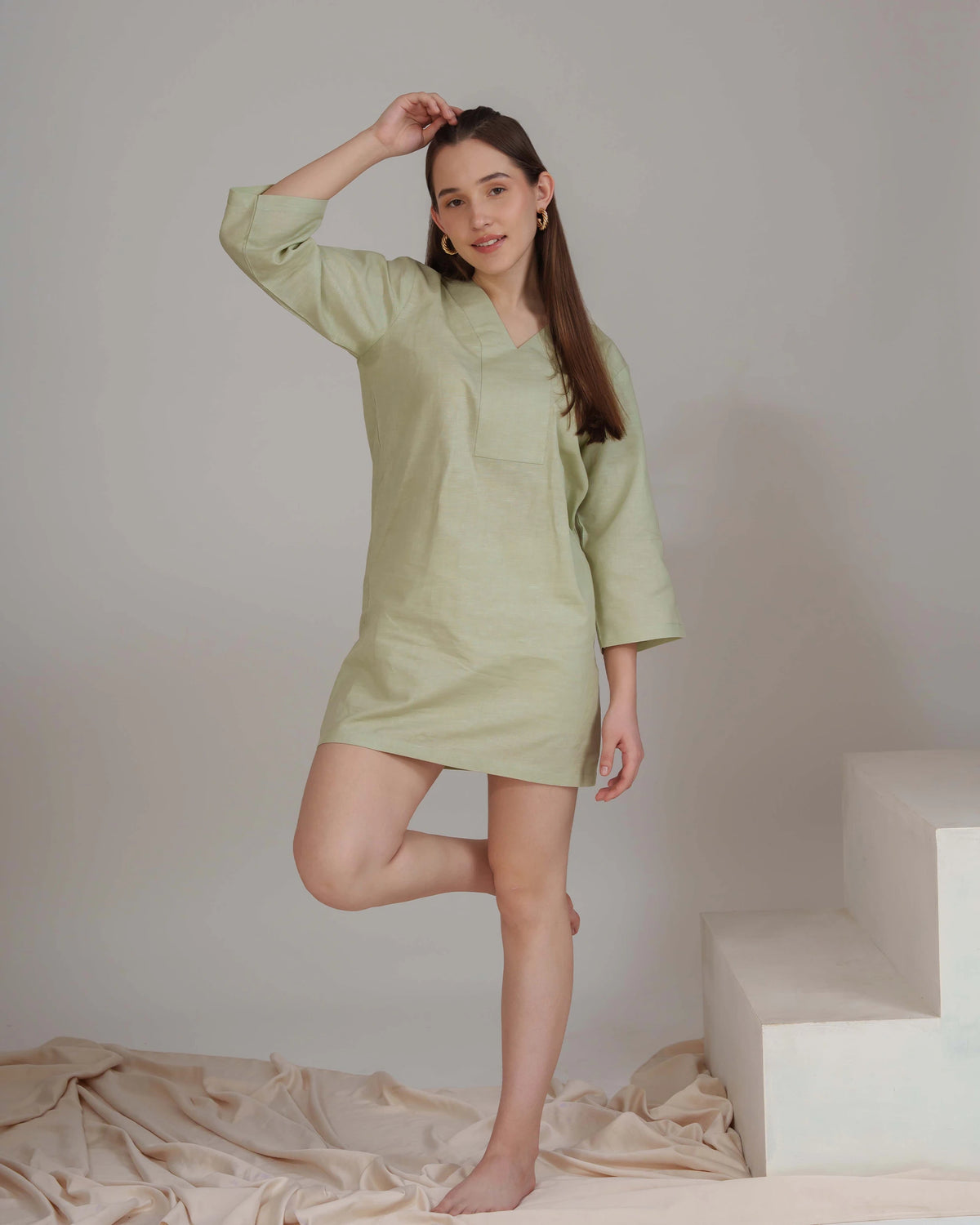 A woman modeling a light sage green linen nightgown or tunic dress with three-quarter sleeves and a chest pocket, standing barefoot on a beige satin sheet against a neutral gray studio background with white steps.