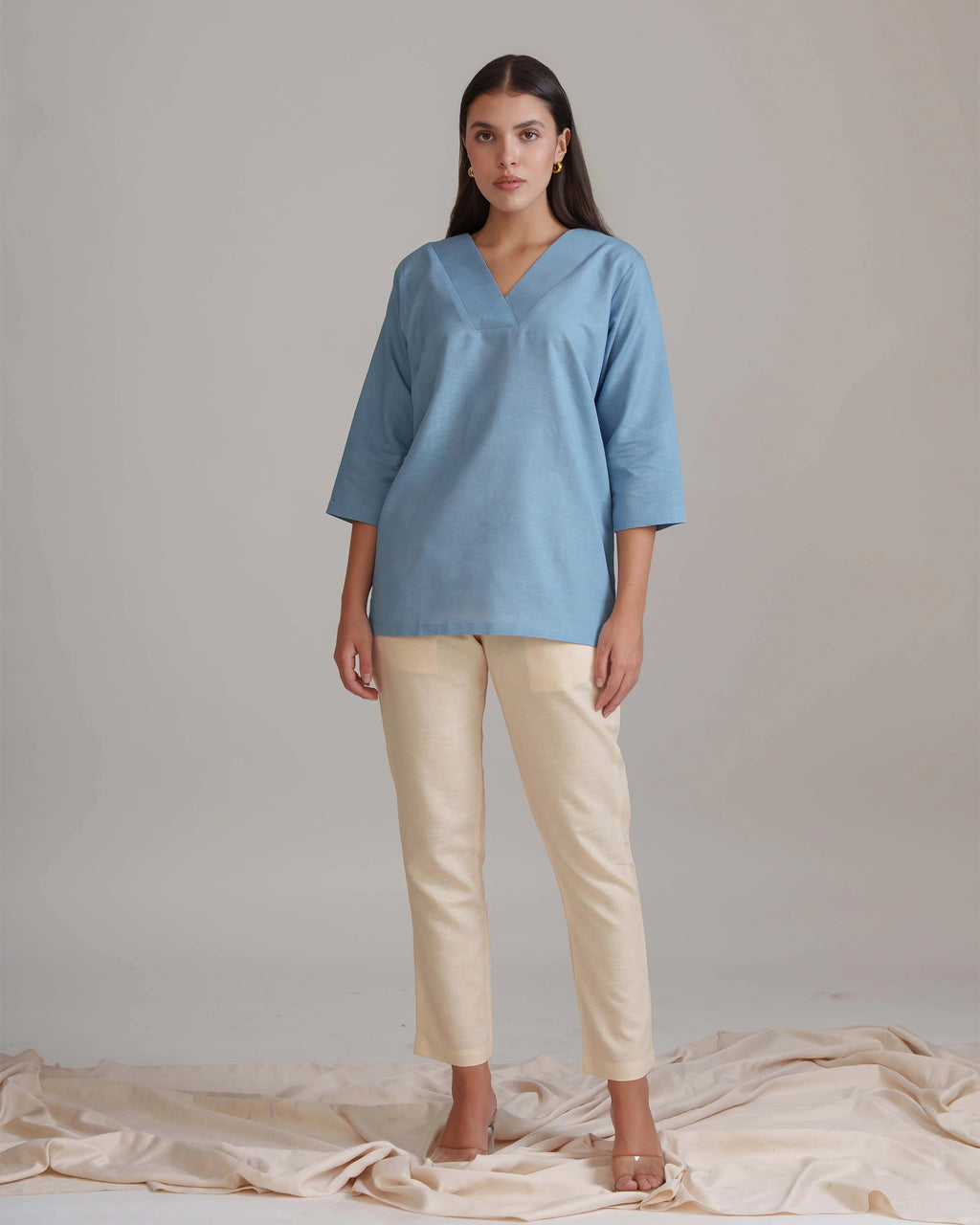 A woman stands against a light beige studio backdrop wearing a loose light blue V-neck top with three-quarter sleeves and beige cropped pants. She is posed on a draped cream fabric surface.
