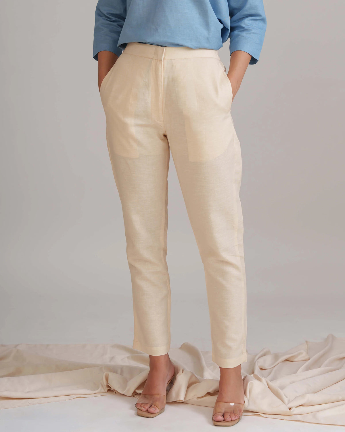A person wearing light beige linen trousers with a blue long-sleeve top and clear heeled sandals, standing against a light gray studio backdrop with draped fabric on the floor.