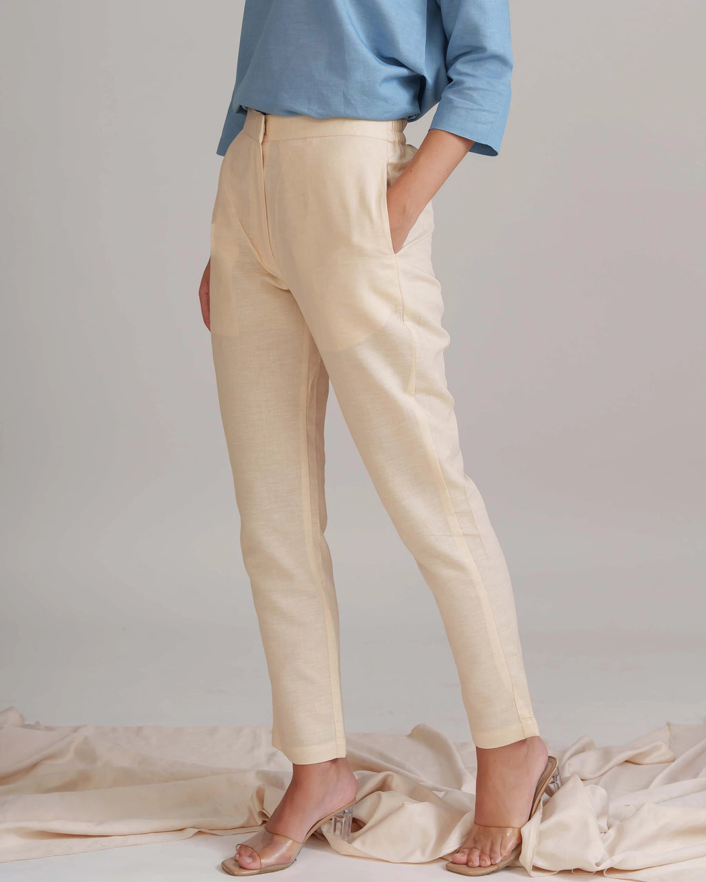 A model wearing a pair of light beige linen trousers with side pockets, paired with a light blue long-sleeve shirt and clear high-heeled sandals, standing against a neutral studio backdrop.