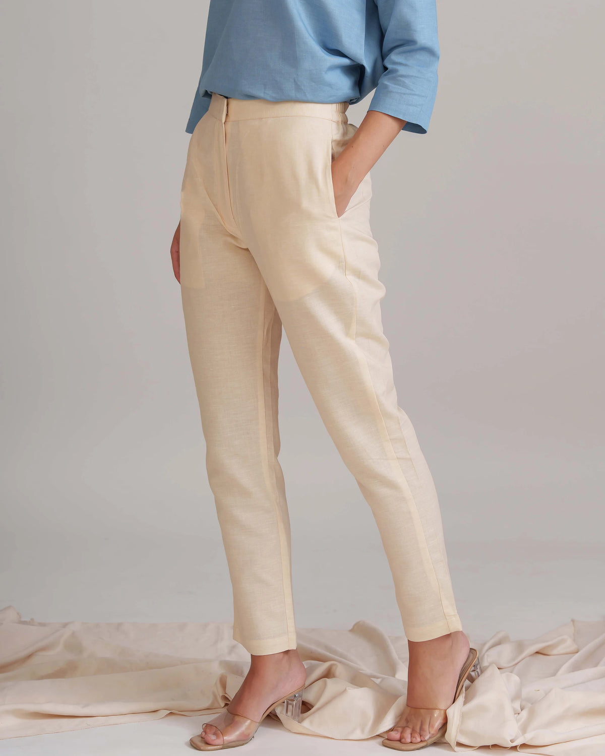 A model wearing a pair of light beige linen trousers with side pockets, paired with a light blue long-sleeve shirt and clear high-heeled sandals, standing against a neutral studio backdrop.