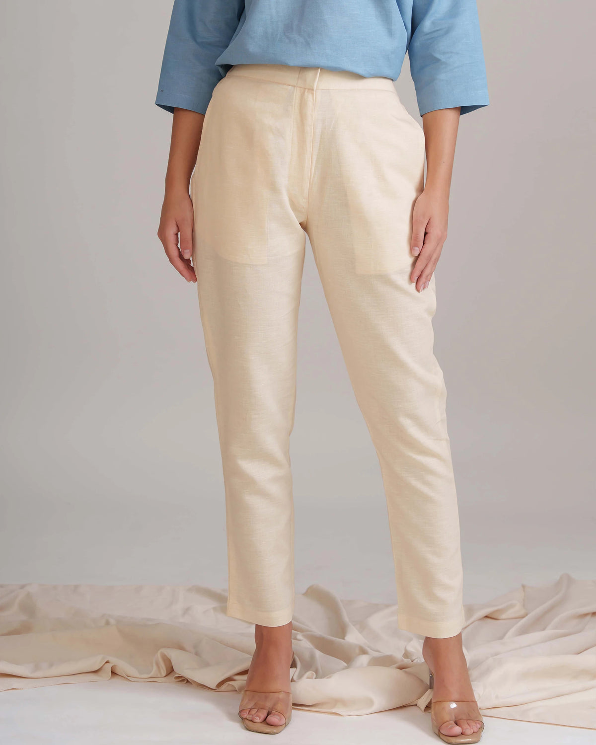 A model wearing a pair of light beige linen trousers with a relaxed straight-leg fit, paired with a light blue top and beige sandals, standing against a neutral studio backdrop with draped fabric at the bottom.