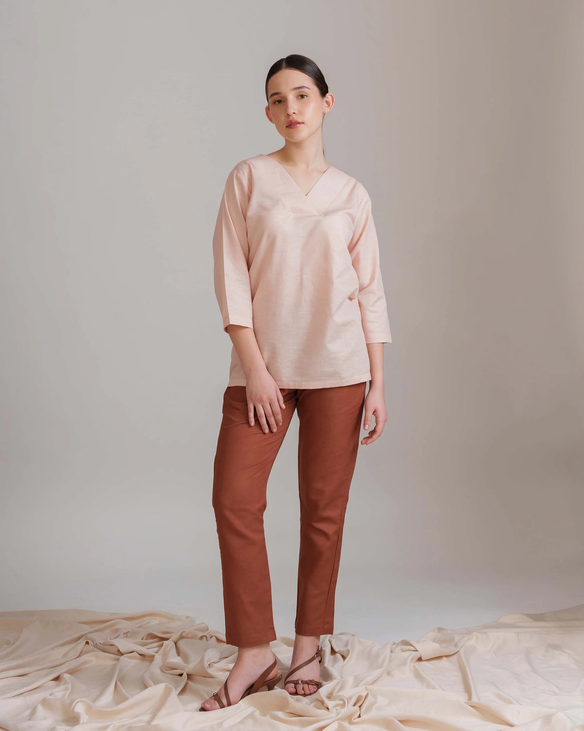 A woman modeling a light pink V-neck three-quarter sleeve top and rust brown cropped pants, standing against a neutral studio backdrop with draped beige fabric at the bottom.