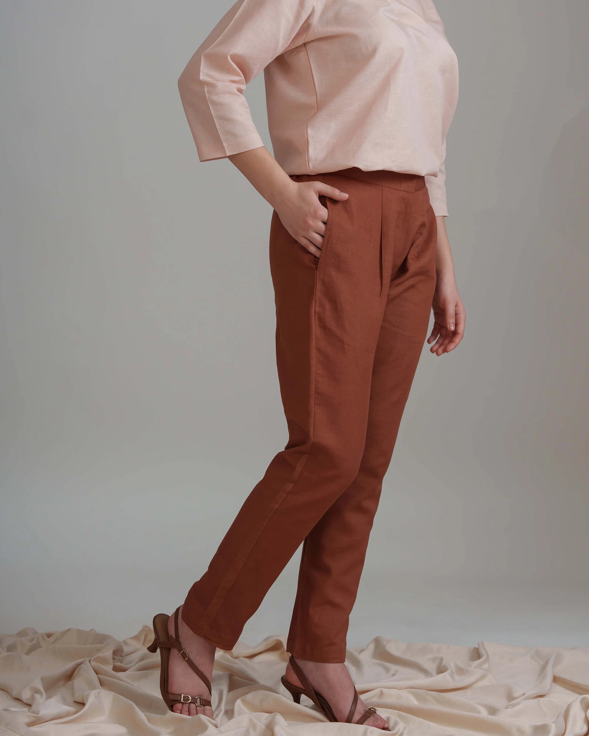 A model wearing rust-brown tailored trousers, a light pink blouse, and brown heeled sandals, standing against a neutral gray studio backdrop with draped beige fabric at the bottom.