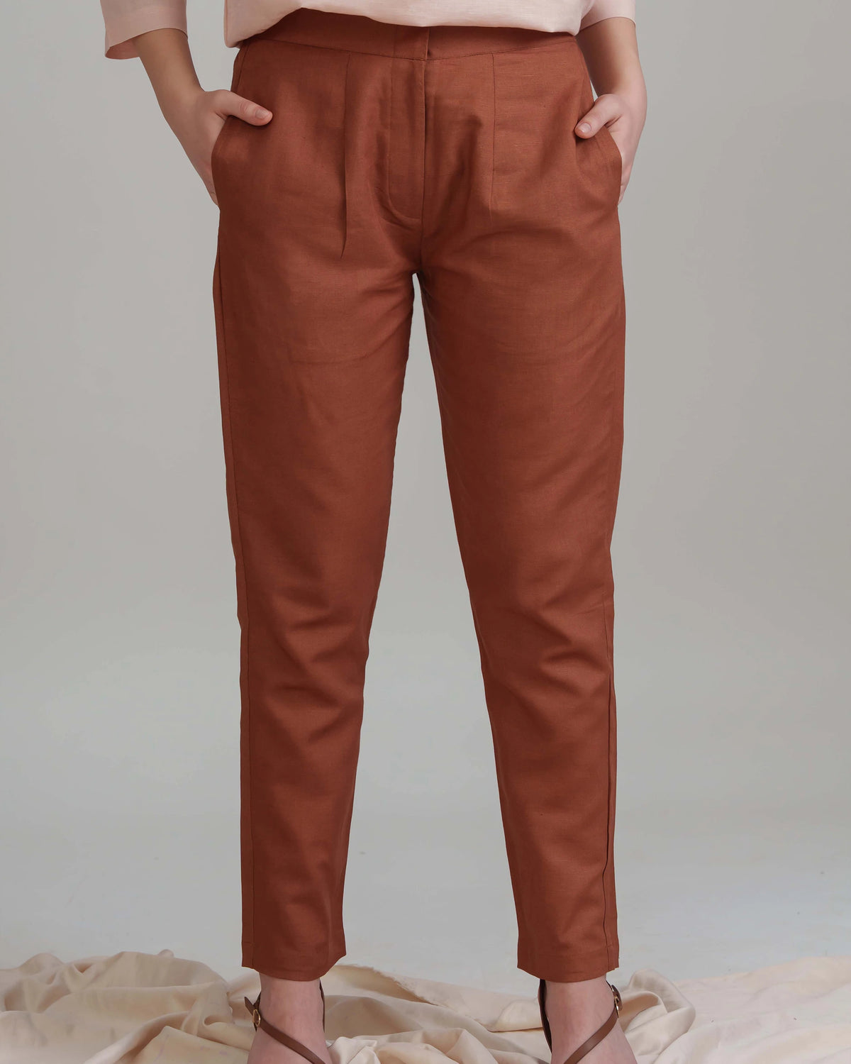A person wearing rust-brown cropped trousers with side pockets, paired with a light pink top and brown strappy heels, standing against a light gray studio background.