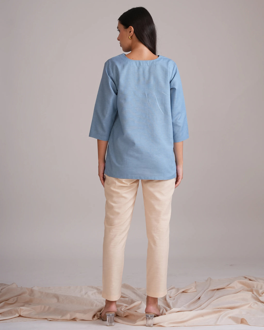 A woman stands with her back to the camera wearing a light blue linen tunic top and beige trousers, standing on a draped beige fabric surface against a neutral studio backdrop.
