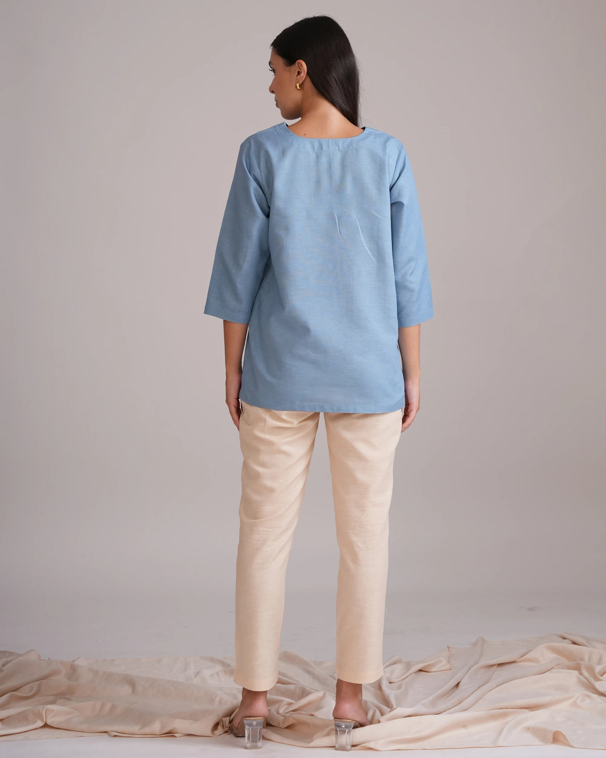 A woman stands with her back to the camera wearing a light blue linen tunic top and beige trousers, standing on a draped beige fabric surface against a neutral studio backdrop.
