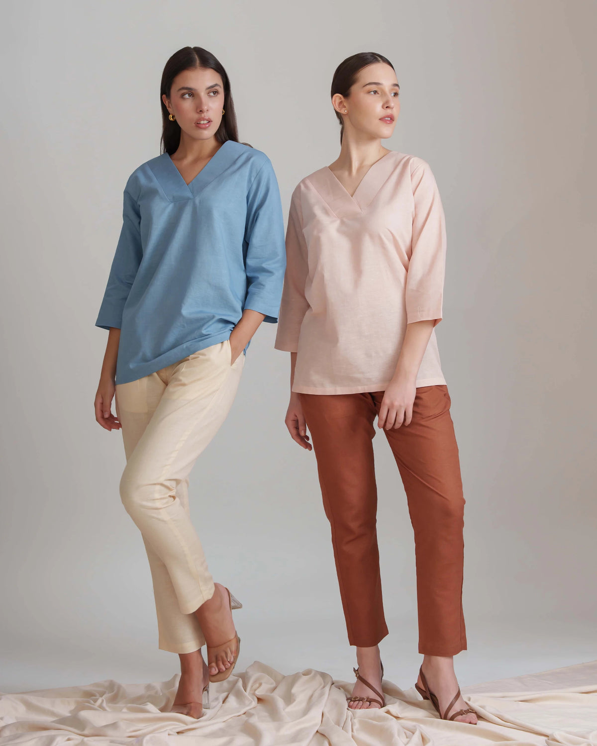 Two women modeling loose V-neck top and trouset set in a studio. The woman on the left wears a light blue top with cream pants, and the woman on the right wears a pale blush pink top with rust brown pants.