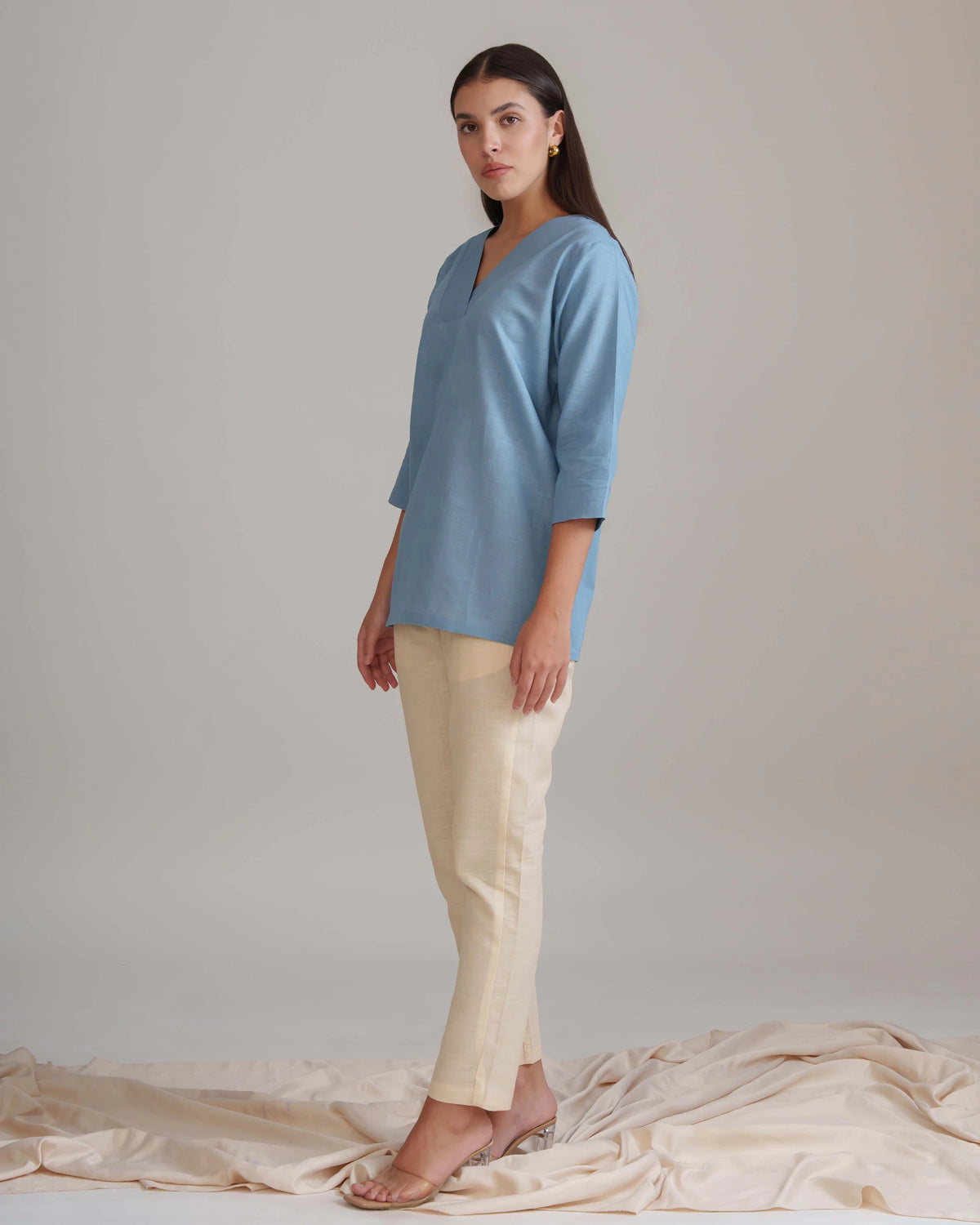 A woman modeling a light blue three-quarter sleeve top and beige trousers, standing against a neutral beige studio backdrop with draped fabric on the floor.