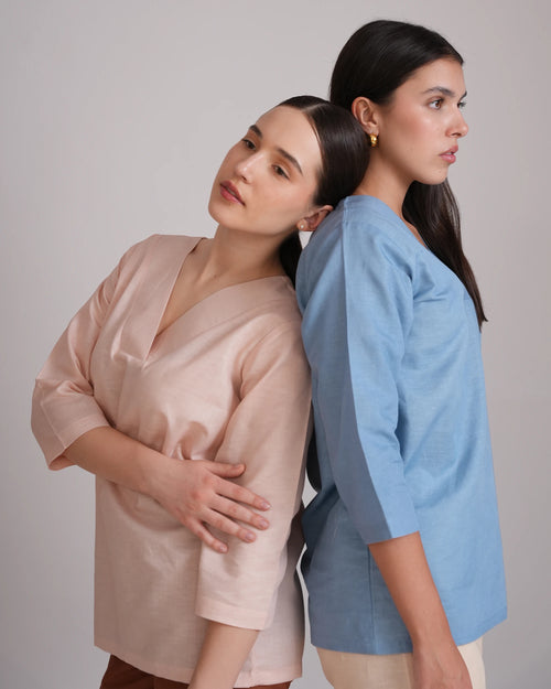 Two women modeling linen-like short-sleeve tops, standing back-to-back against a plain light gray studio background. One top is a soft blush pink and the other is a dusty light blue.