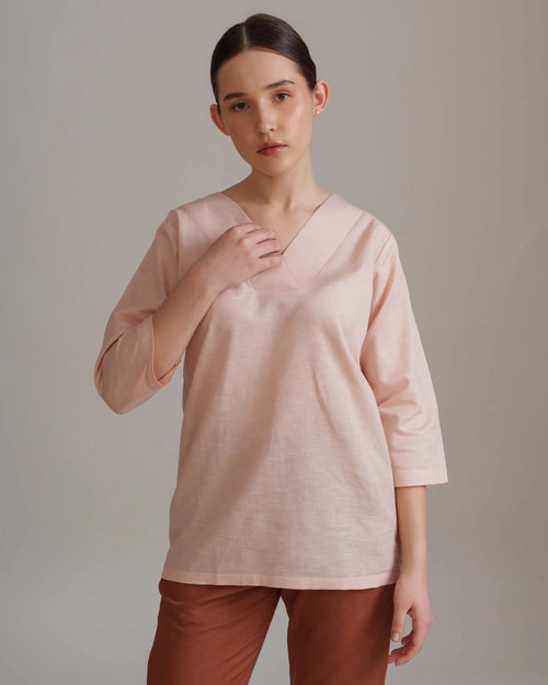 A woman modeling a light pink linen-blend top with a V-neck and elbow-length sleeves, paired with rust-brown pants, against a neutral gray studio background.