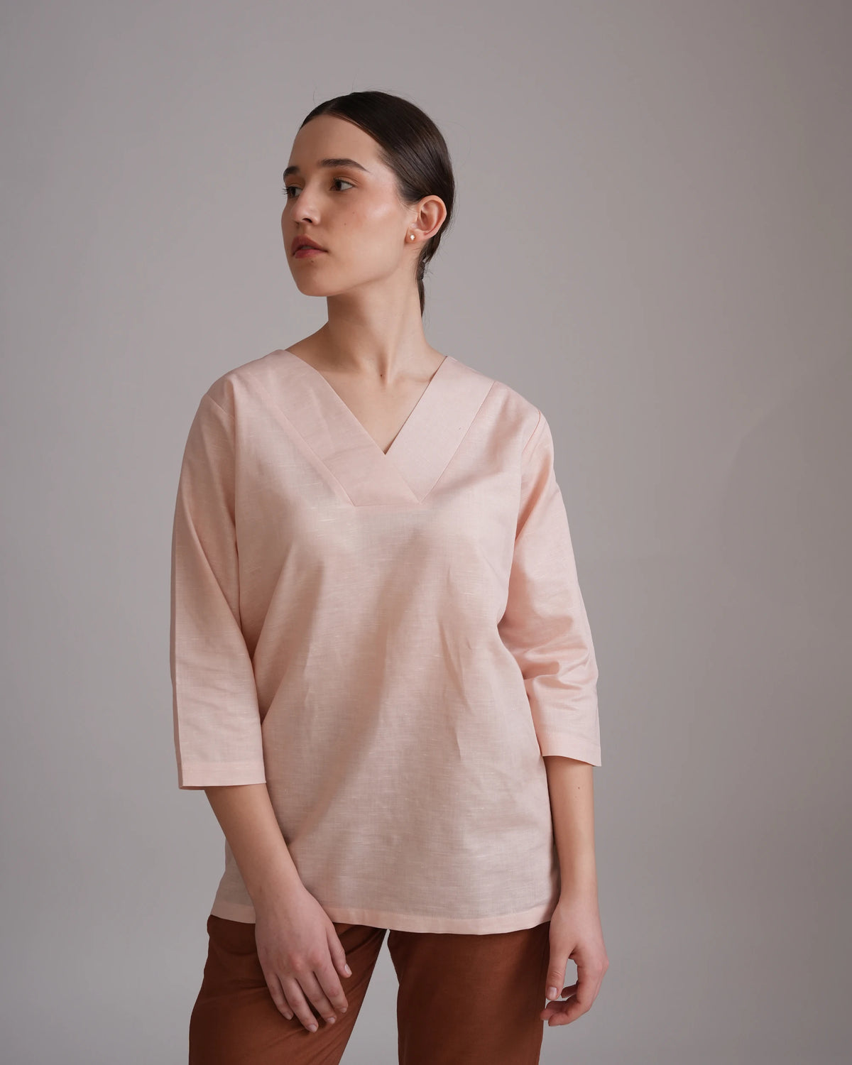 A woman wearing a light blush pink wrap-style top with three-quarter sleeves and brown pants, standing against a neutral gray studio background.
