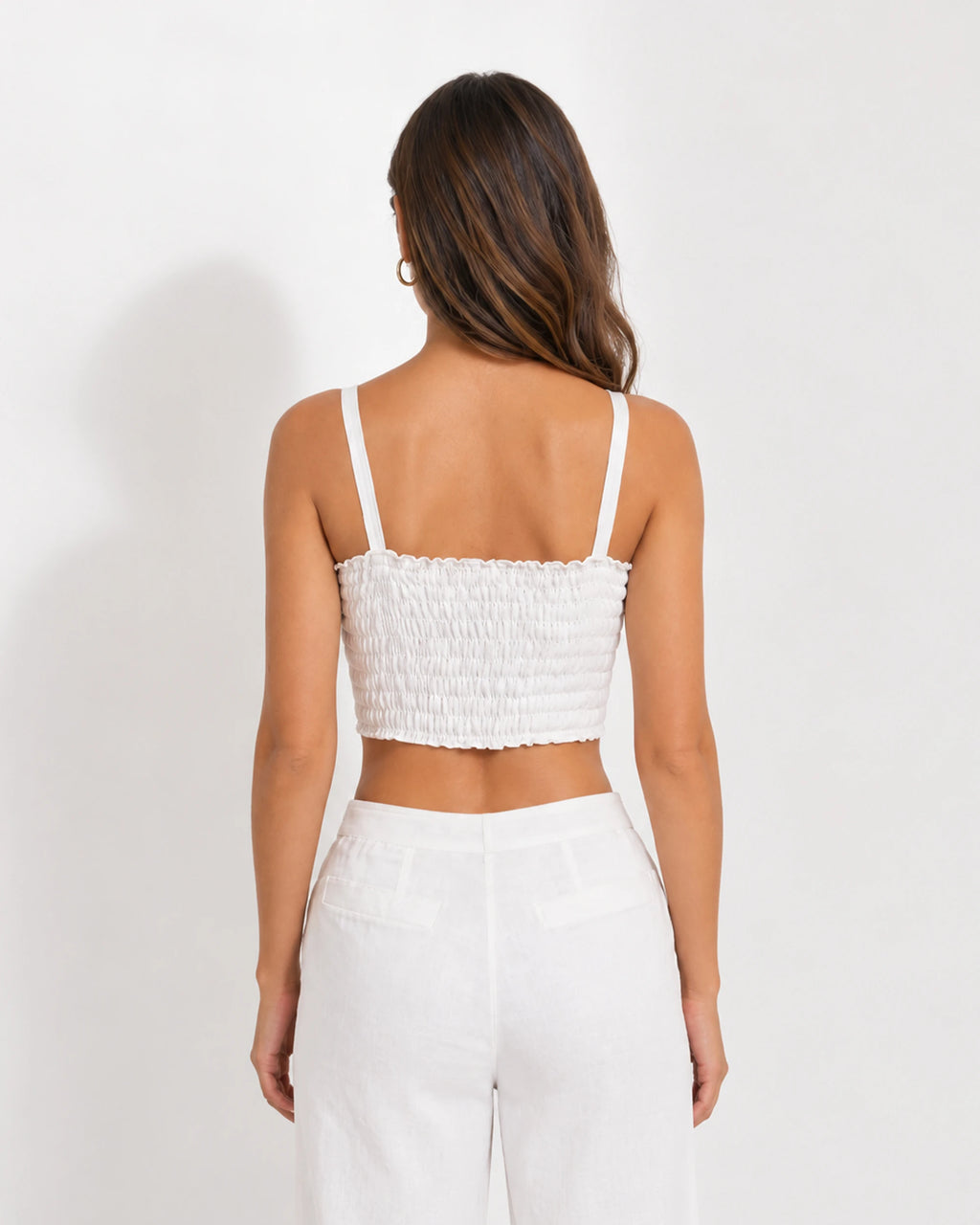 A woman shown from the back wearing a white smocked crop top with thin spaghetti straps and white high-waisted pants against a plain white studio background.