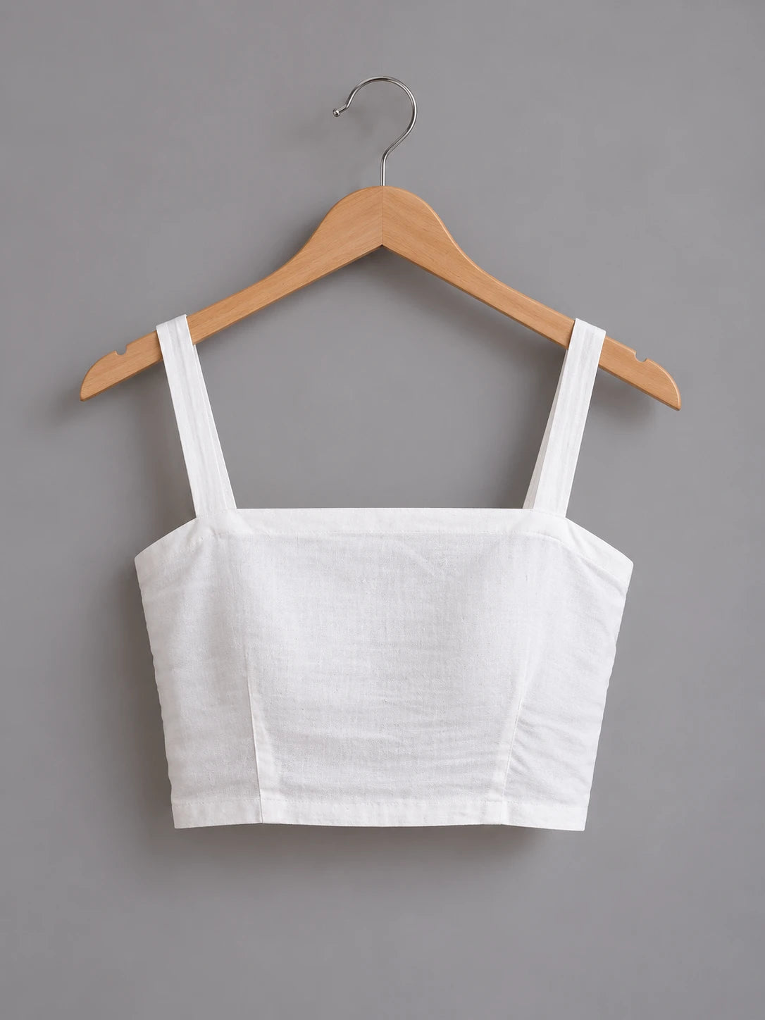 A white linen crop top with square neckline and wide shoulder straps, hanging on a wooden hanger against a solid gray background.