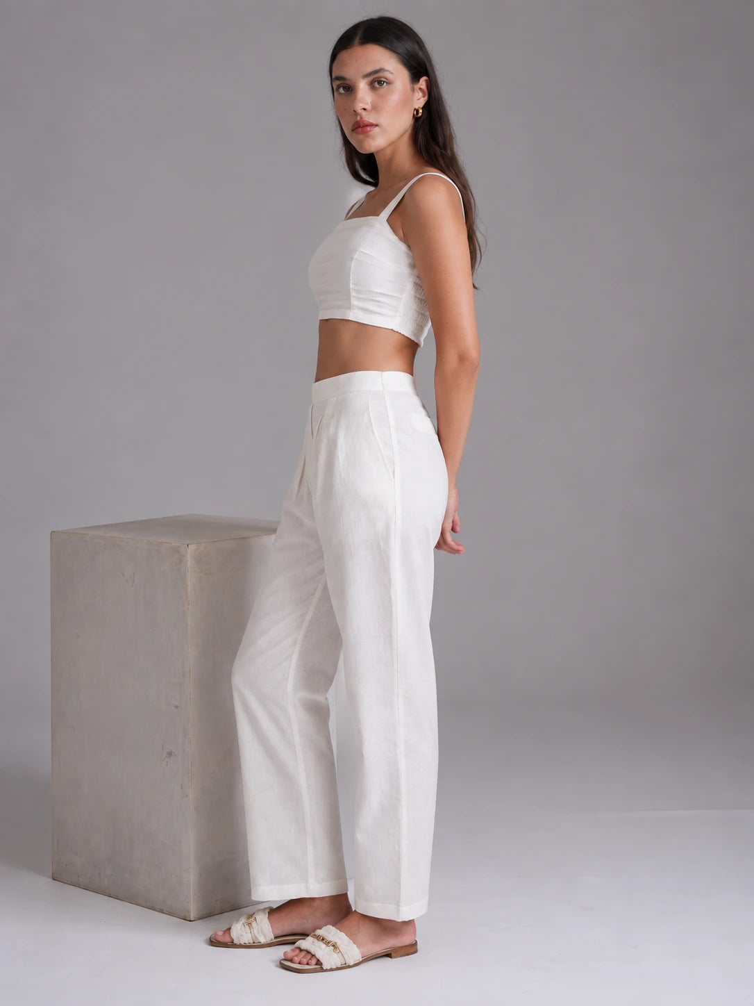 A woman modeling a white two-piece outfit consisting of a sleeveless crop top and matching wide-leg trousers, standing next to a concrete block against a neutral gray studio background.