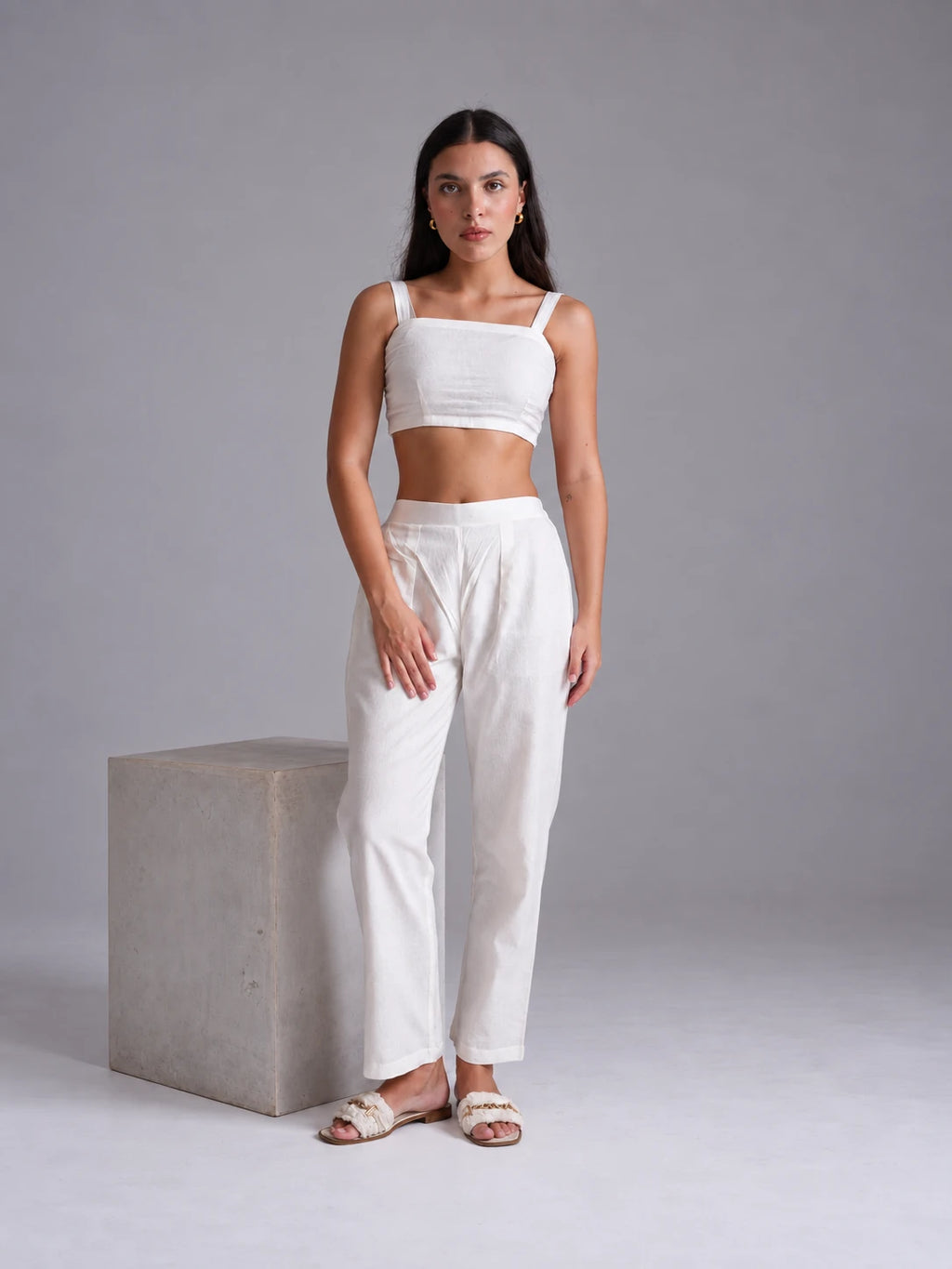 A woman modeling a white two-piece outfit consisting of a square-neck crop top and matching high-waisted wide-leg trousers, standing next to a concrete block against a neutral gray studio background.
