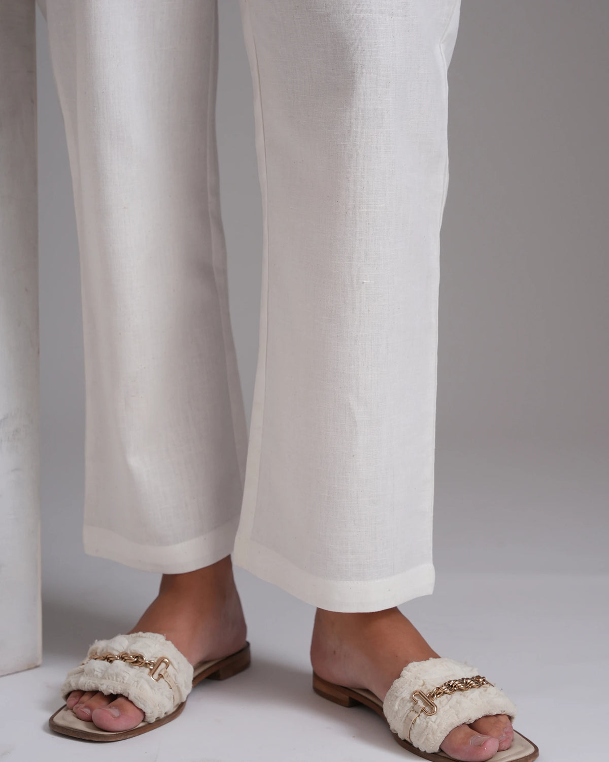 A close-up of a person's lower legs wearing white linen wide-leg trousers and fluffy off-white slide sandals with gold-tone chain details, set against a neutral gray background.
