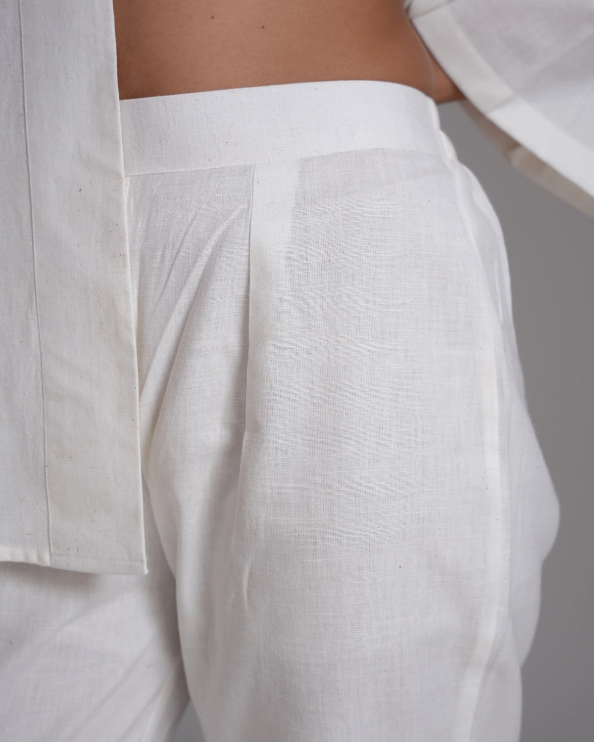 Close-up of a person wearing white linen trousers, showing the side and back waist with a pleated detail, paired with a matching white shirt.