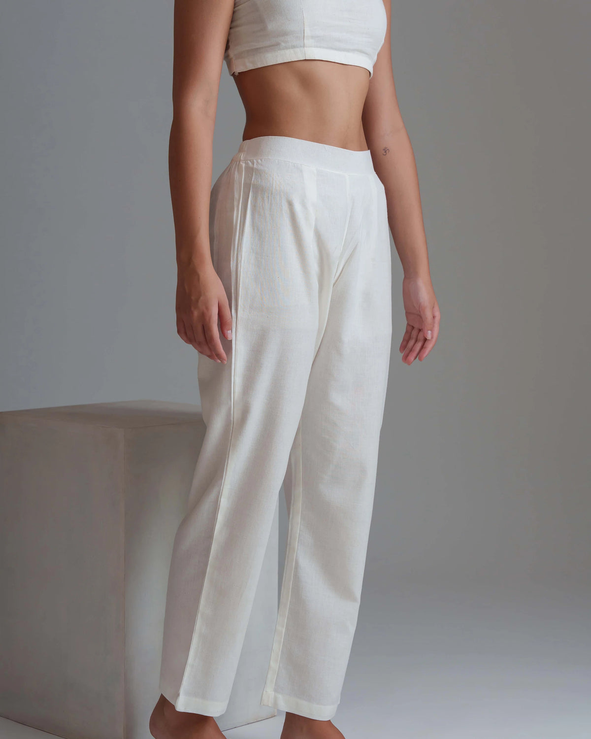 A model wearing high-waisted, wide-leg white linen pants, shown from the waist down in a studio setting with a neutral gray background.