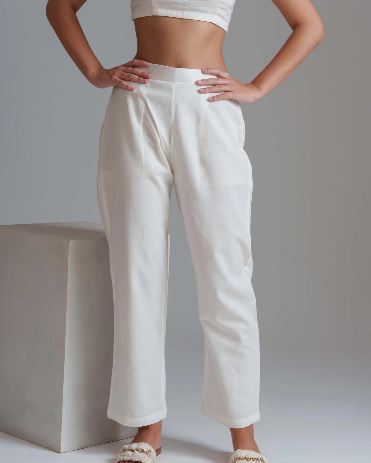 A model wearing high-waisted, wide-leg white trousers with a relaxed straight-to-cropped fit, paired with a white crop top and beige woven sandals, standing next to a minimalist concrete block against a neutral gray background.