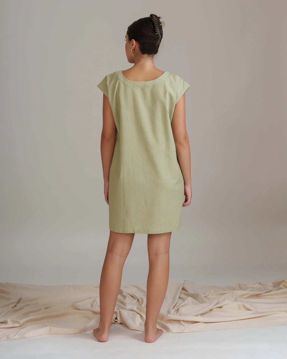 A woman stands with her back to the camera wearing a light sage green short-sleeve tunic dress. She is standing on a white floor with beige fabric draped around her feet, against a neutral beige background.