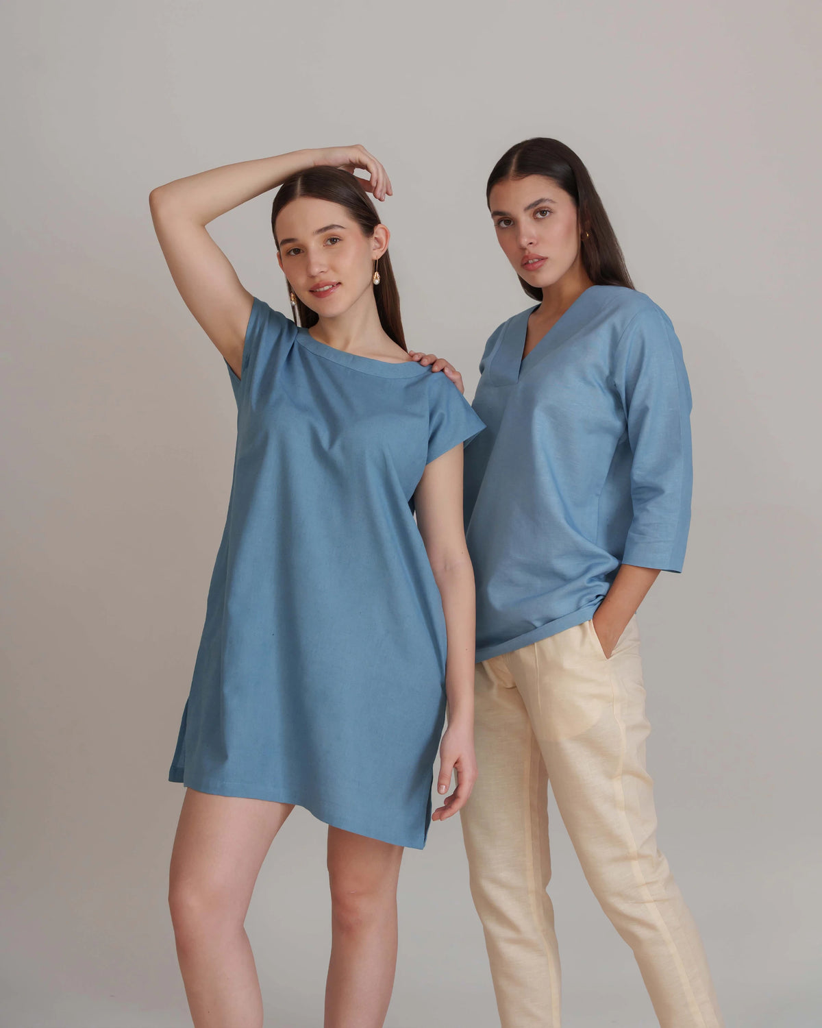 Two women modeling blue tops in a studio setting against a light gray background. The woman on the left wears an oversized short-sleeve blue tunic-style top, and the woman on the right wears a matching blue V-neck top with three-quarter sleeves paired with light beige pants.