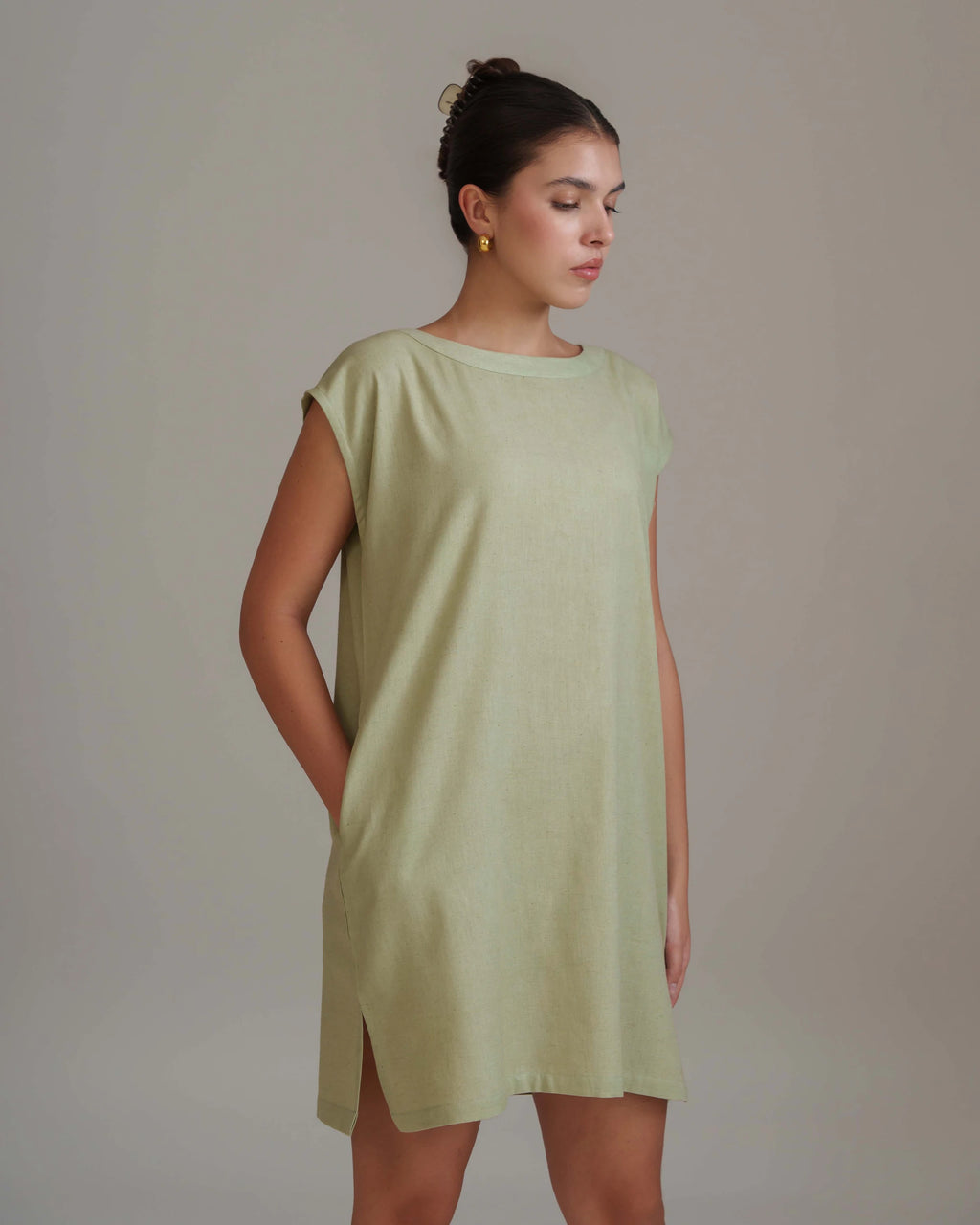 A woman wearing a light sage green linen tunic dress with a relaxed, oversized fit and short cap sleeves, standing against a neutral beige studio background.