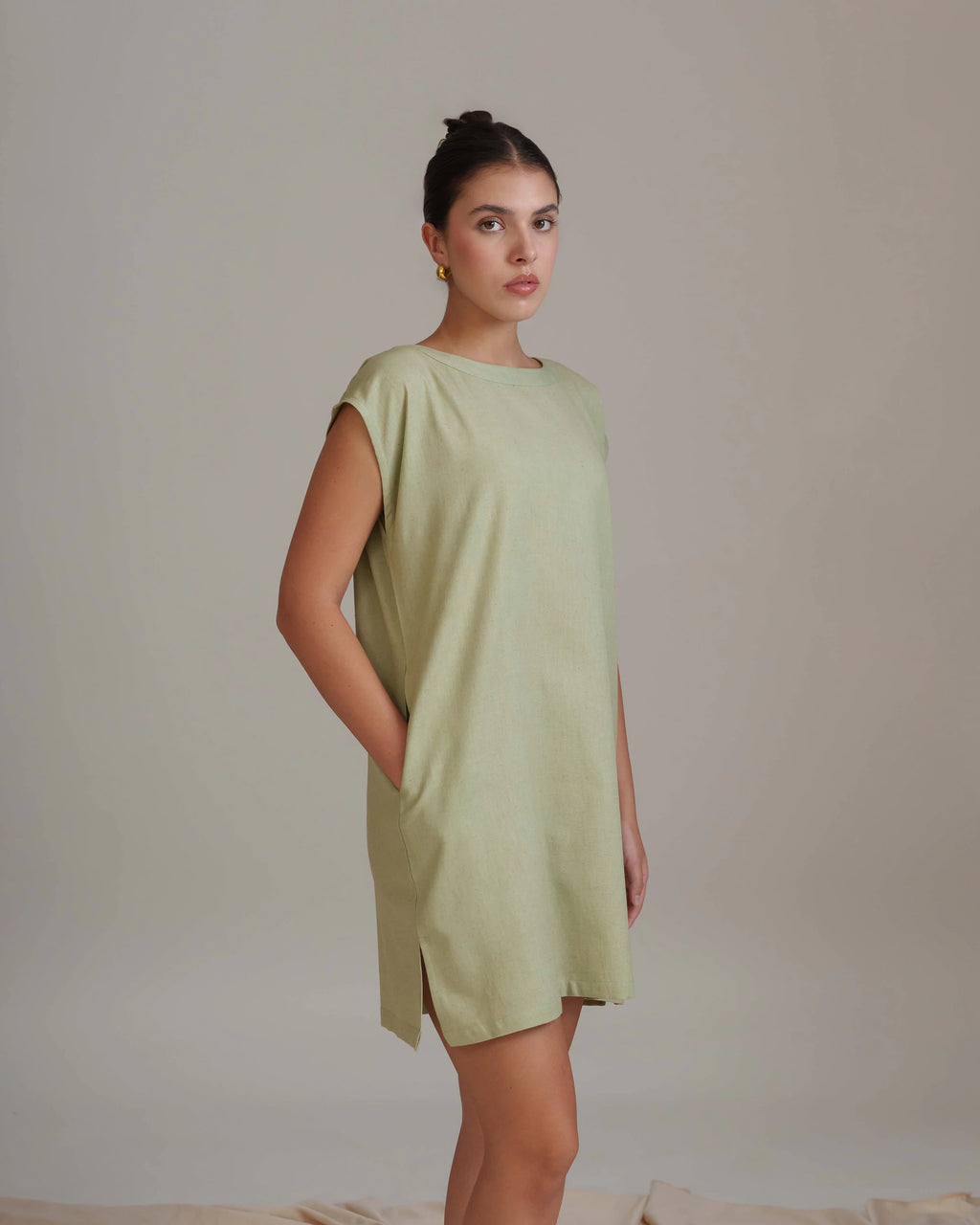A woman modeling a light sage green sleeveless tunic dress with side slits, standing against a neutral beige studio backdrop.