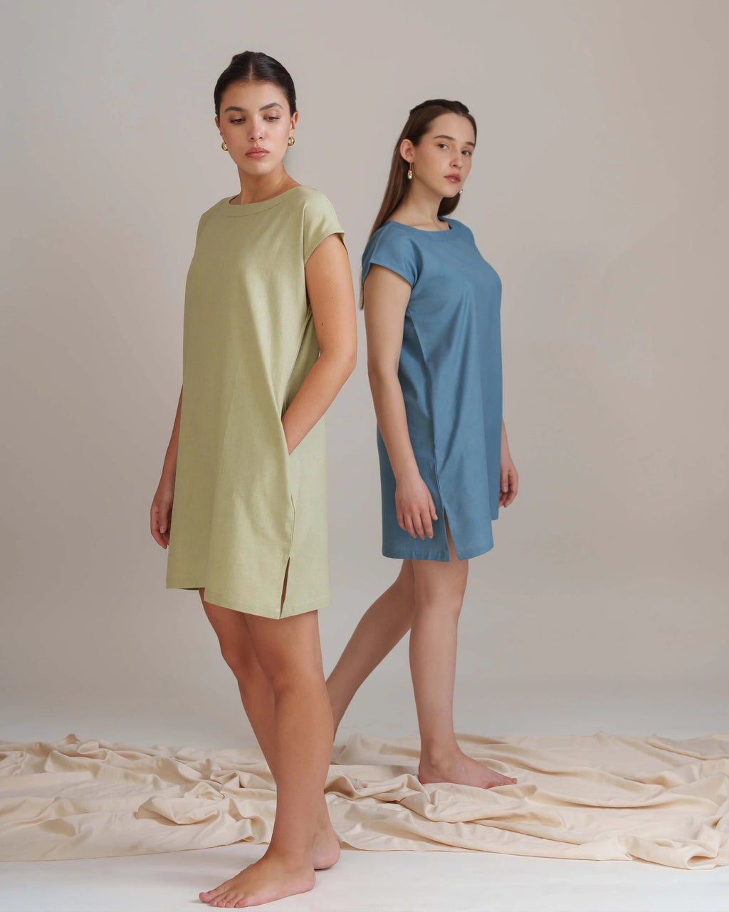 Two women modeling short-sleeve shift dresses in a studio setting. The woman in front wears a light sage green dress, and the woman behind wears a dusty blue dress.