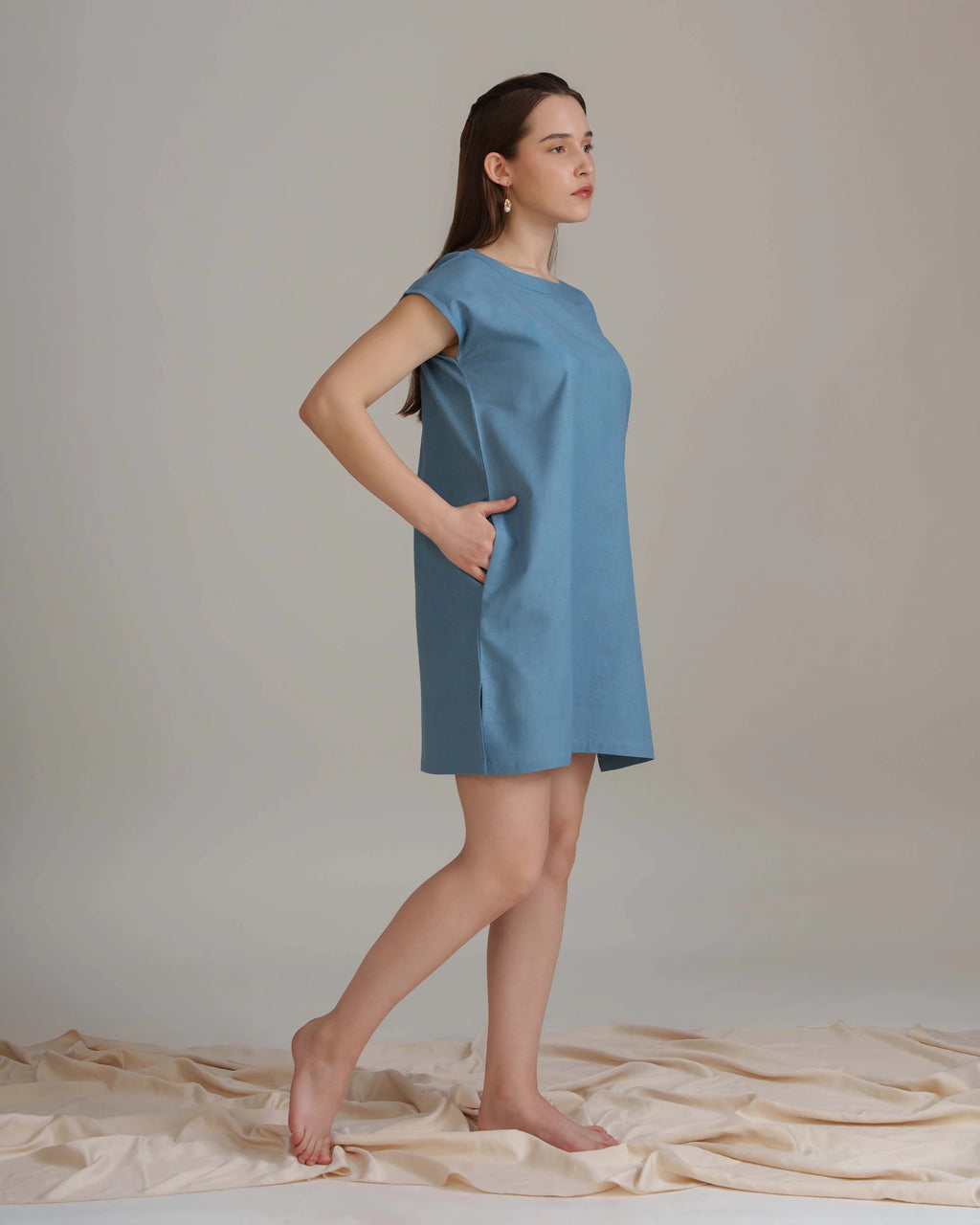A woman modeling a short-sleeve, knee-length blue dress with side pockets, standing barefoot on a draped beige fabric against a neutral studio backdrop.