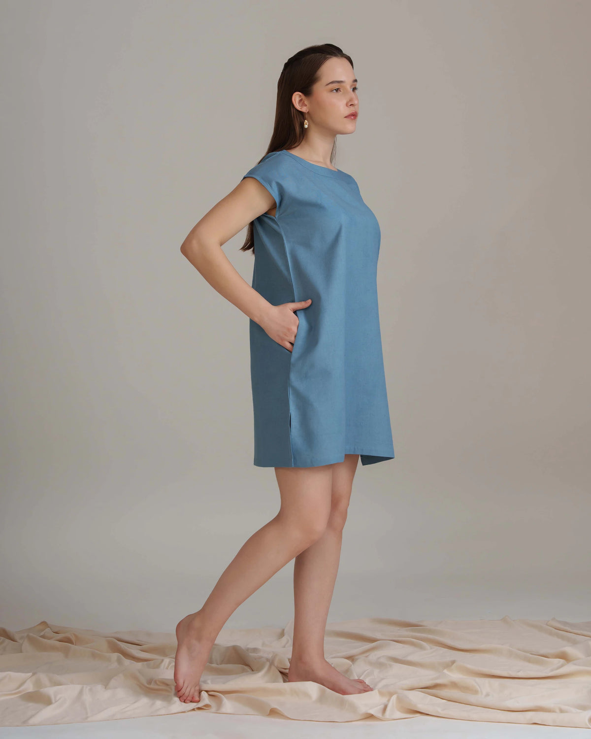A woman modeling a short-sleeve, knee-length blue dress with side pockets, standing barefoot on a draped beige fabric against a neutral studio backdrop.