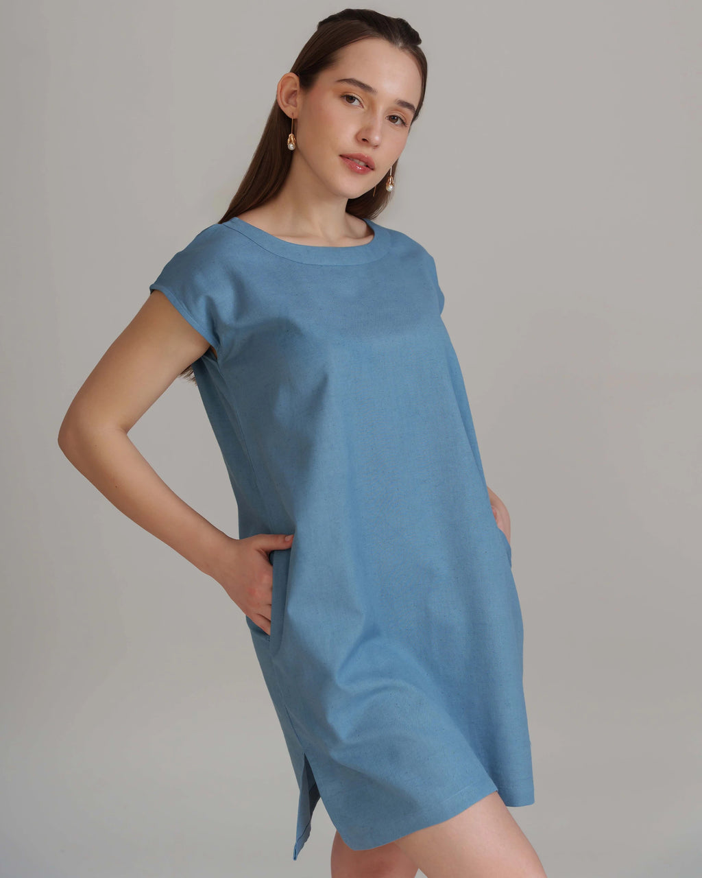 A woman modeling a short-sleeve, loose-fitting blue dress with side pockets against a plain light gray studio background.