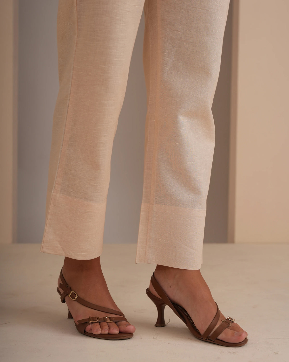 Close-up of a person's legs wearing light beige linen trousers and brown strappy heeled sandals with a buckle detail, set against a minimalist beige background.